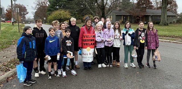 A group of children and adults pose outdoors holding a cylindrical food drive banner.