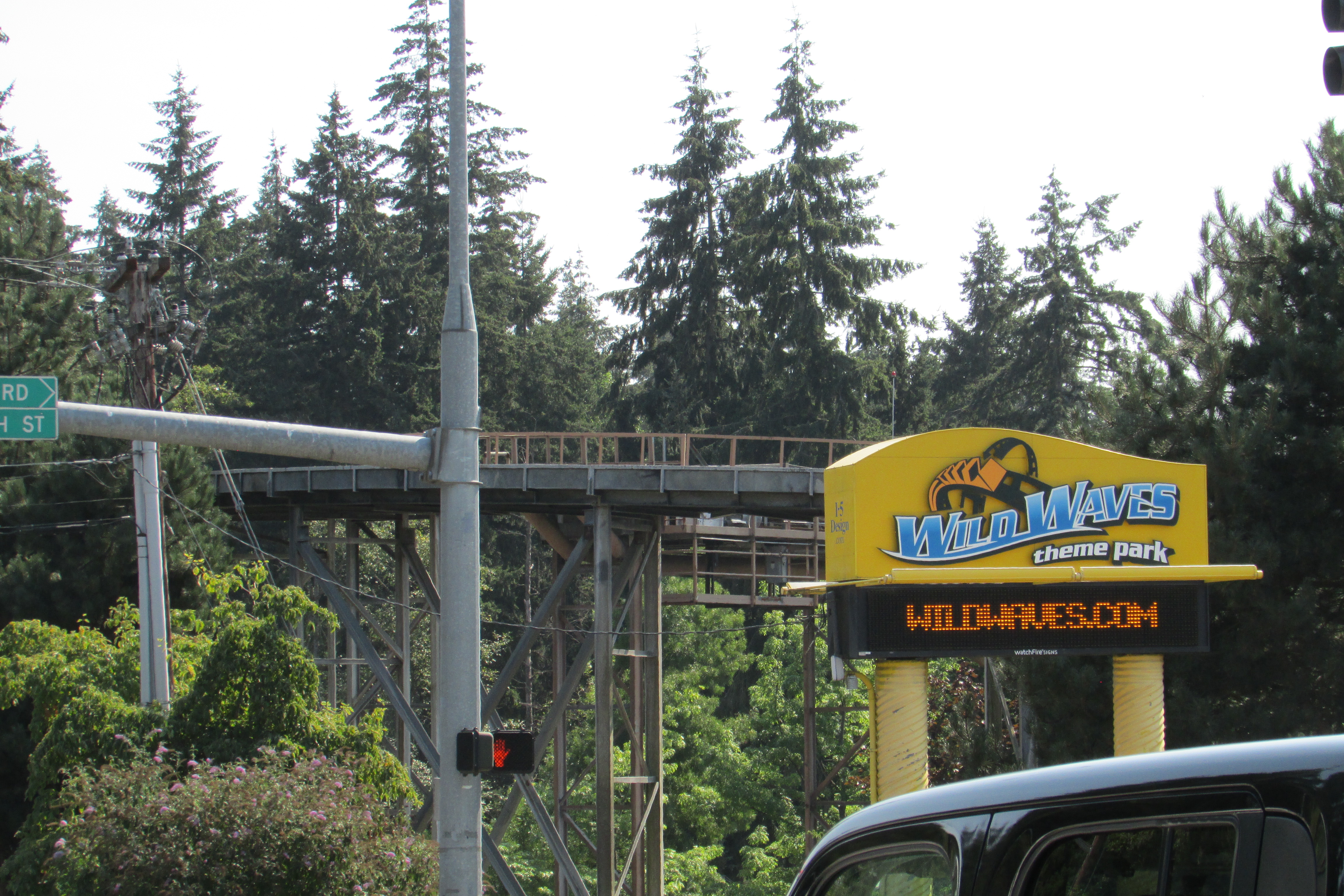 Roadside sign reading Wild Waves Theme and Water Park with an elevated ride structure and tall evergreen trees behind it.