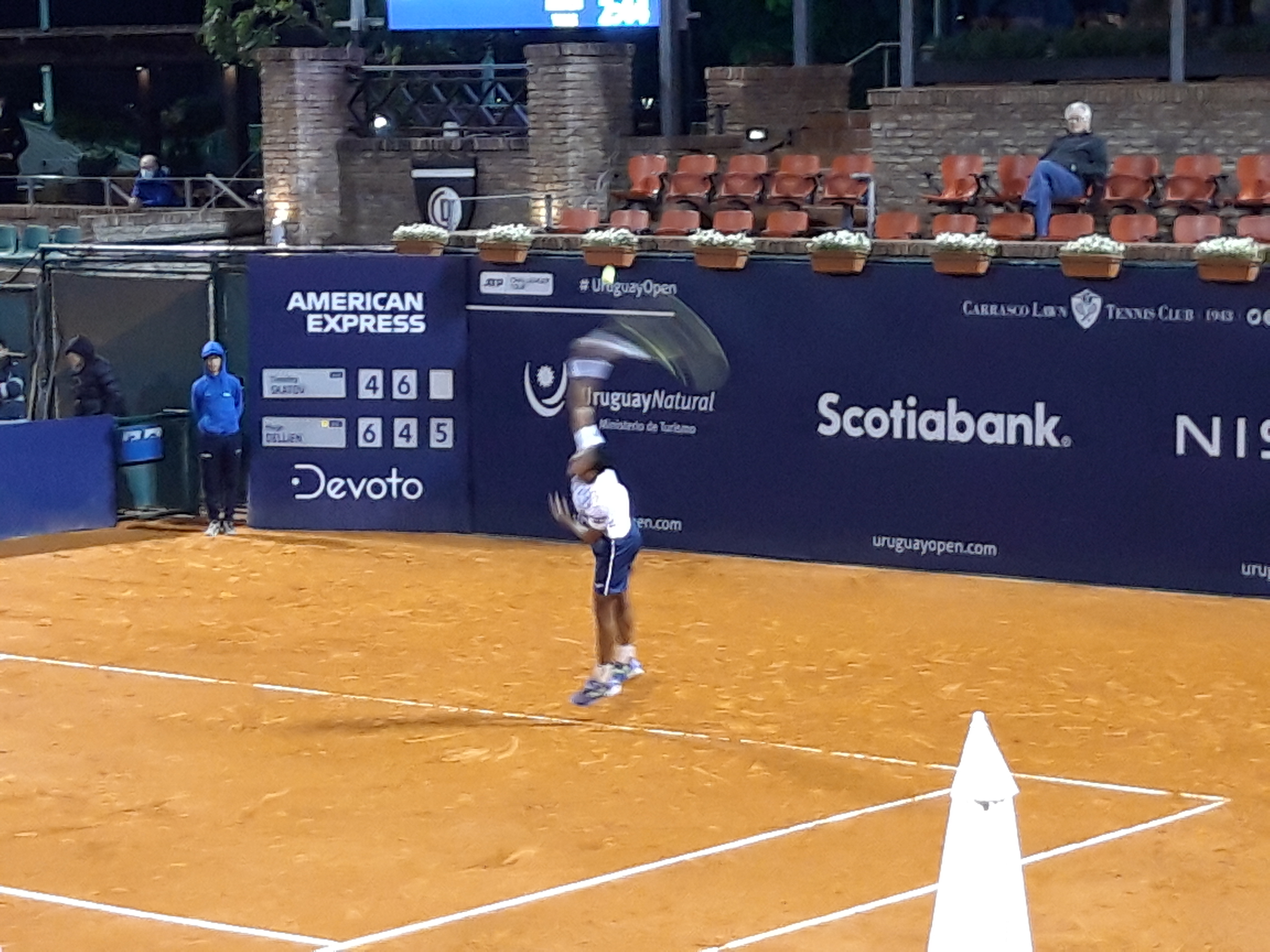Tennis player mid-serve on a clay court with sponsor banners and spectators behind.