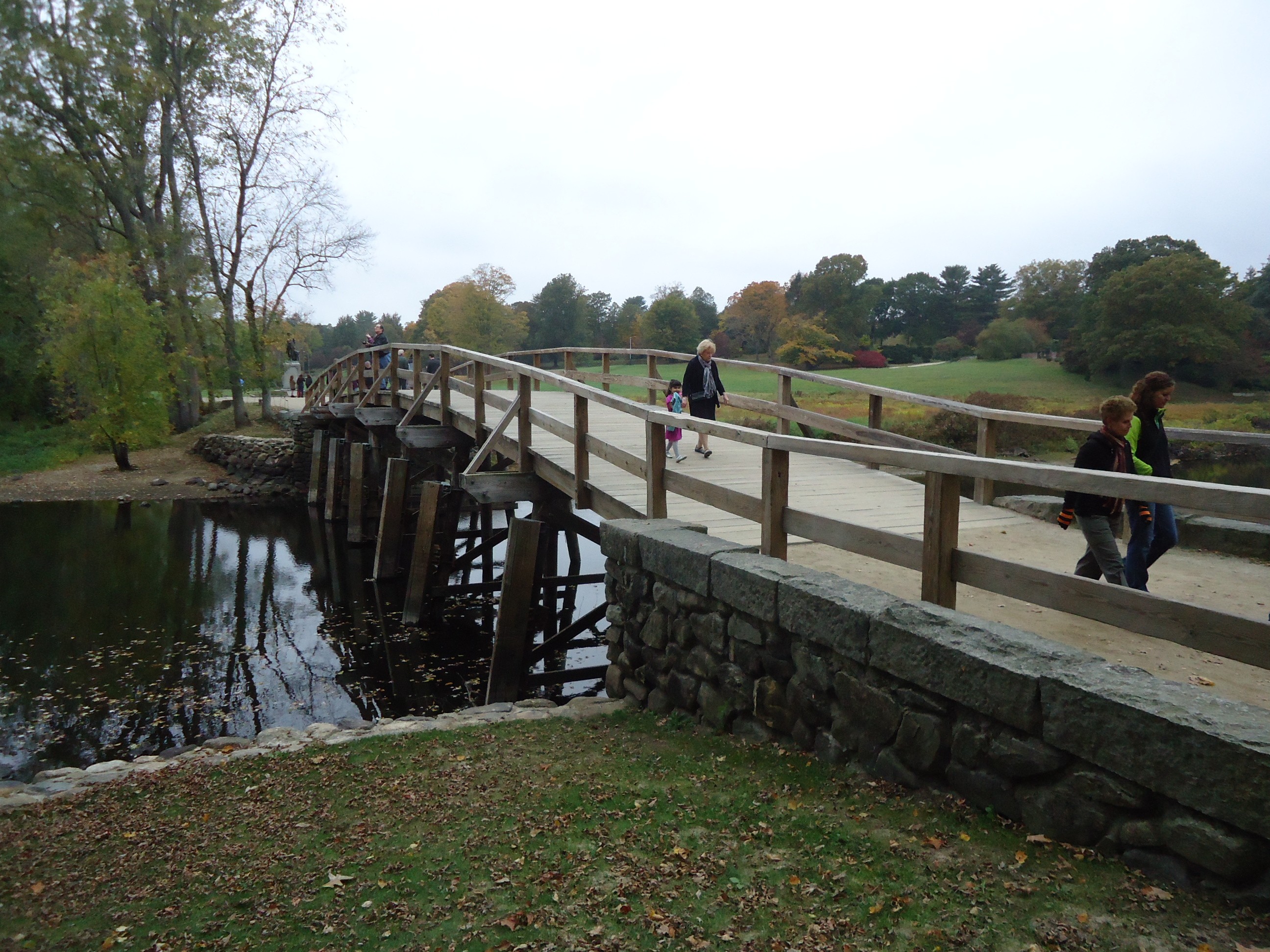 Visitors walking across a wooden pedestrian bridge over a river surrounded by autumn foliage.
