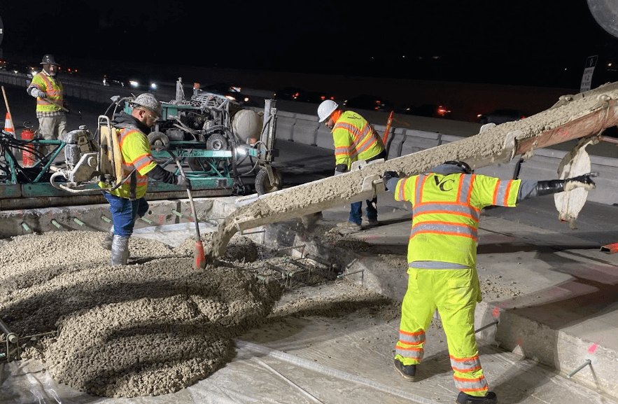 Construction workers in high-visibility gear pour concrete on a freeway at night under artificial lighting.