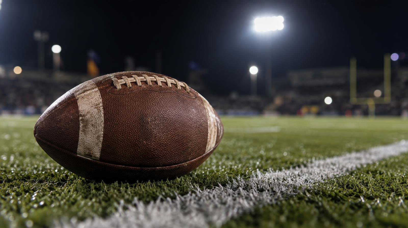 An American football resting on a green grass field beside a white yard line under stadium lights.