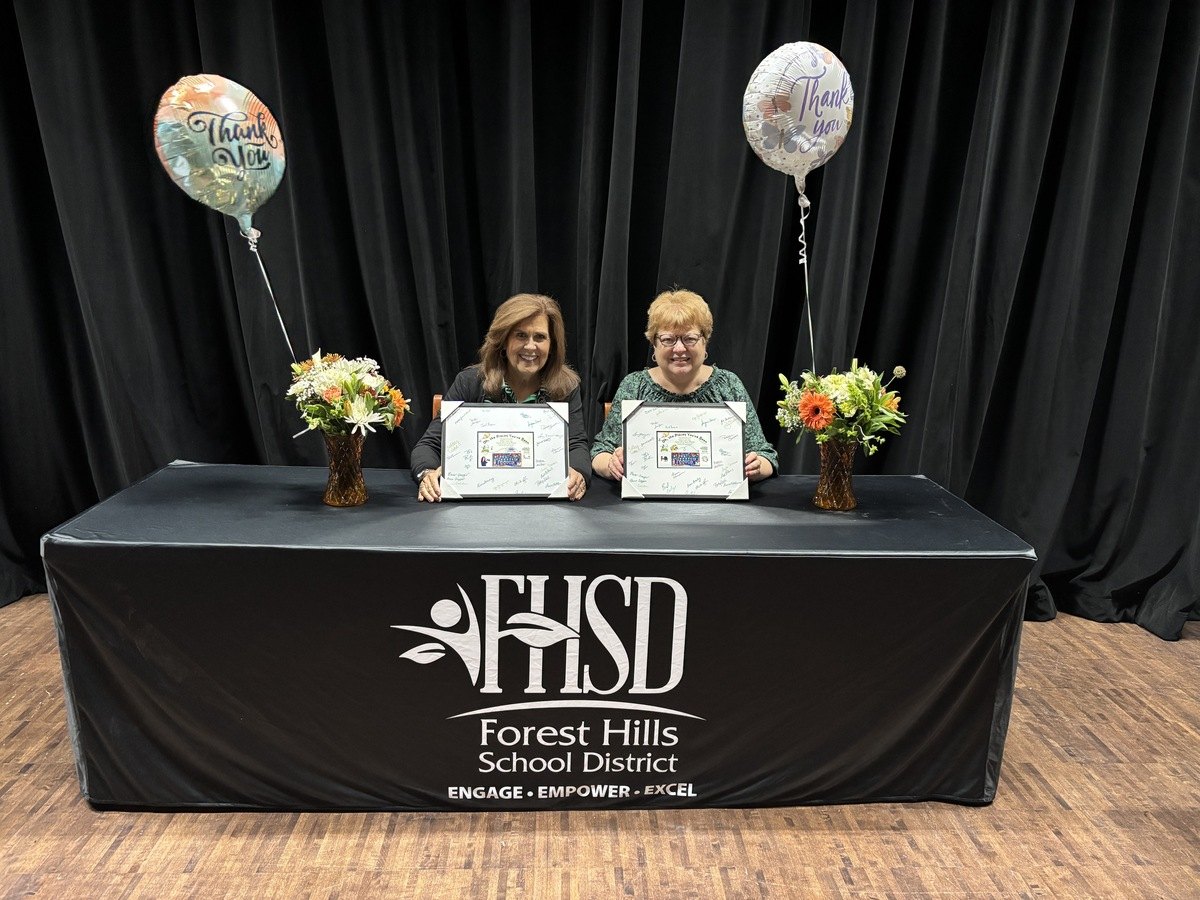 Two women seated at a table holding framed certificates, with floral arrangements and thank-you balloons.