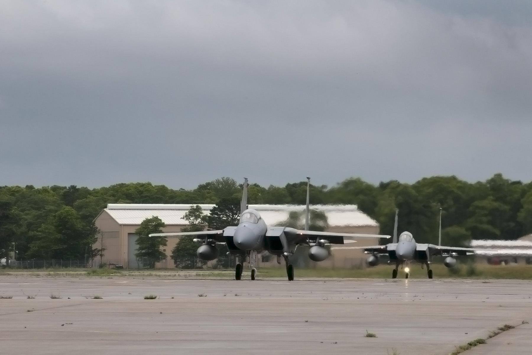 Two F-15 Eagle jets taxiing on a runway apron toward military hangars.