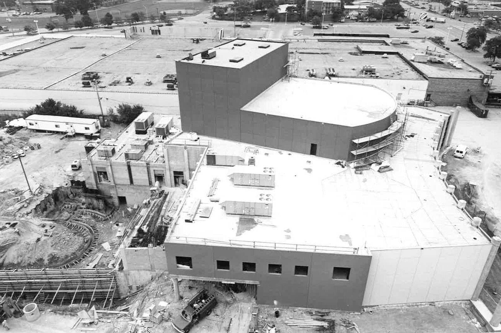 Black-and-white aerial photo of a performing arts center under construction with a curved theater structure and surrounding parking areas.