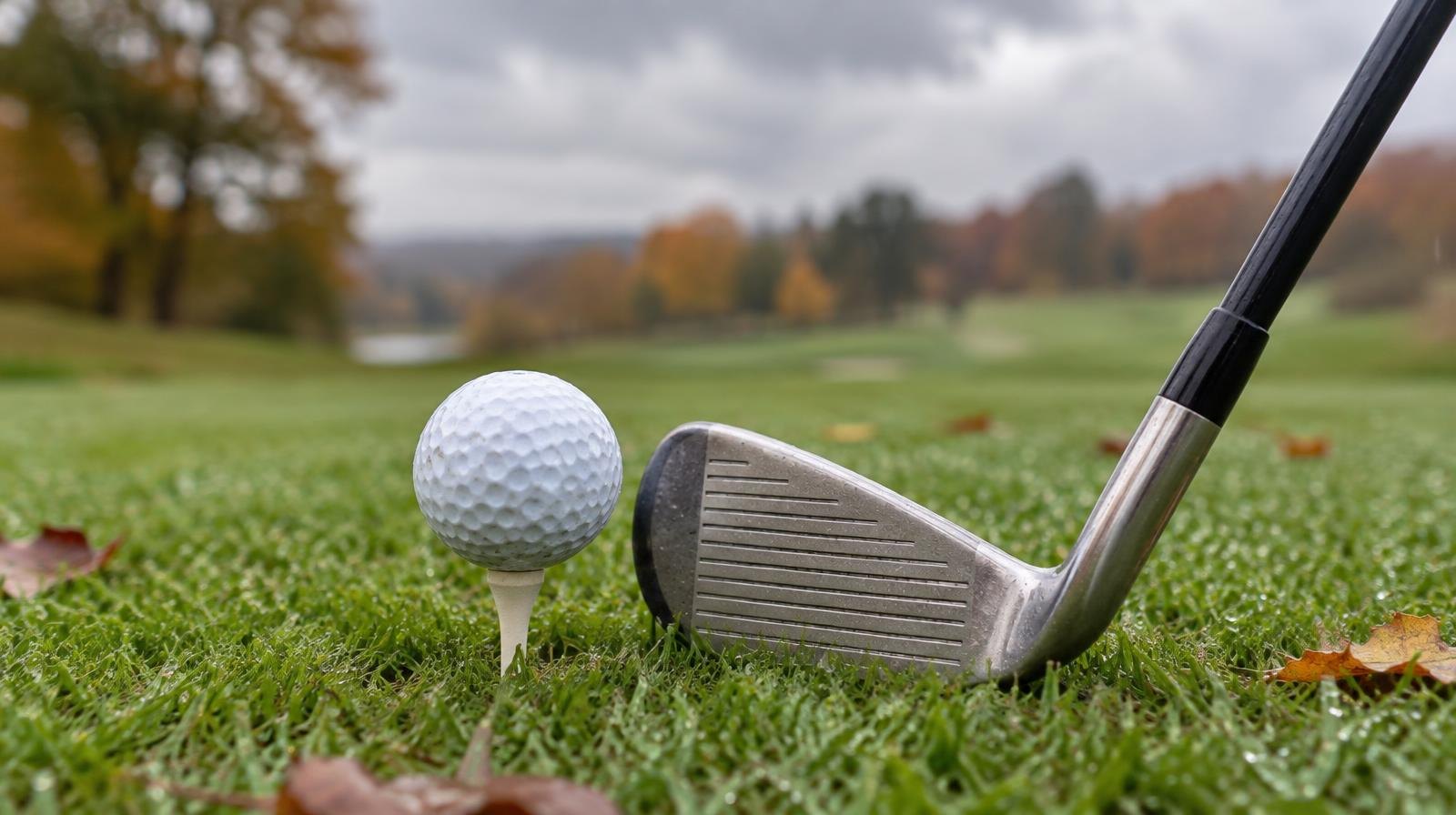 A golf ball on a tee beside an iron club on a wet, misty fairway.