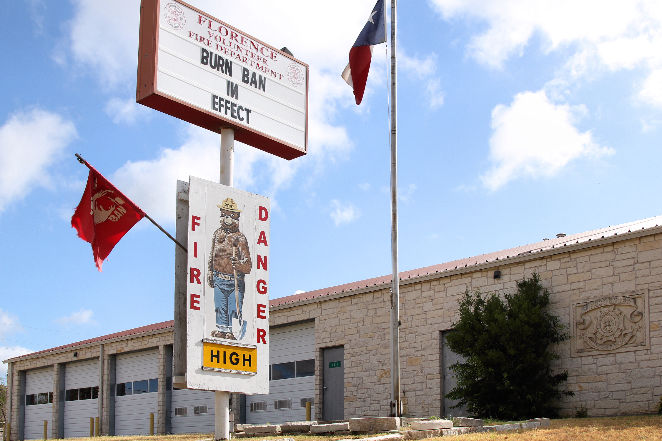 Fire station sign reading 'Burn Ban In Effect' with a fire danger rating below and a Texas flag nearby.