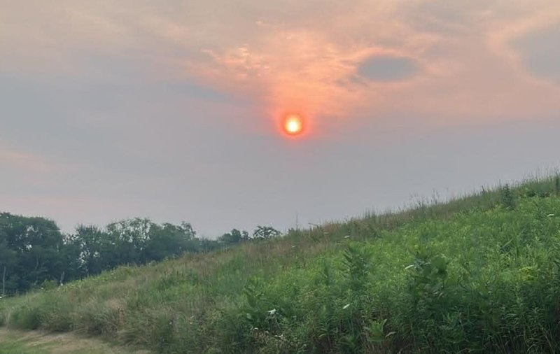 Red-orange sun partially obscured by haze above a green grassy hillside under a smoky sky.