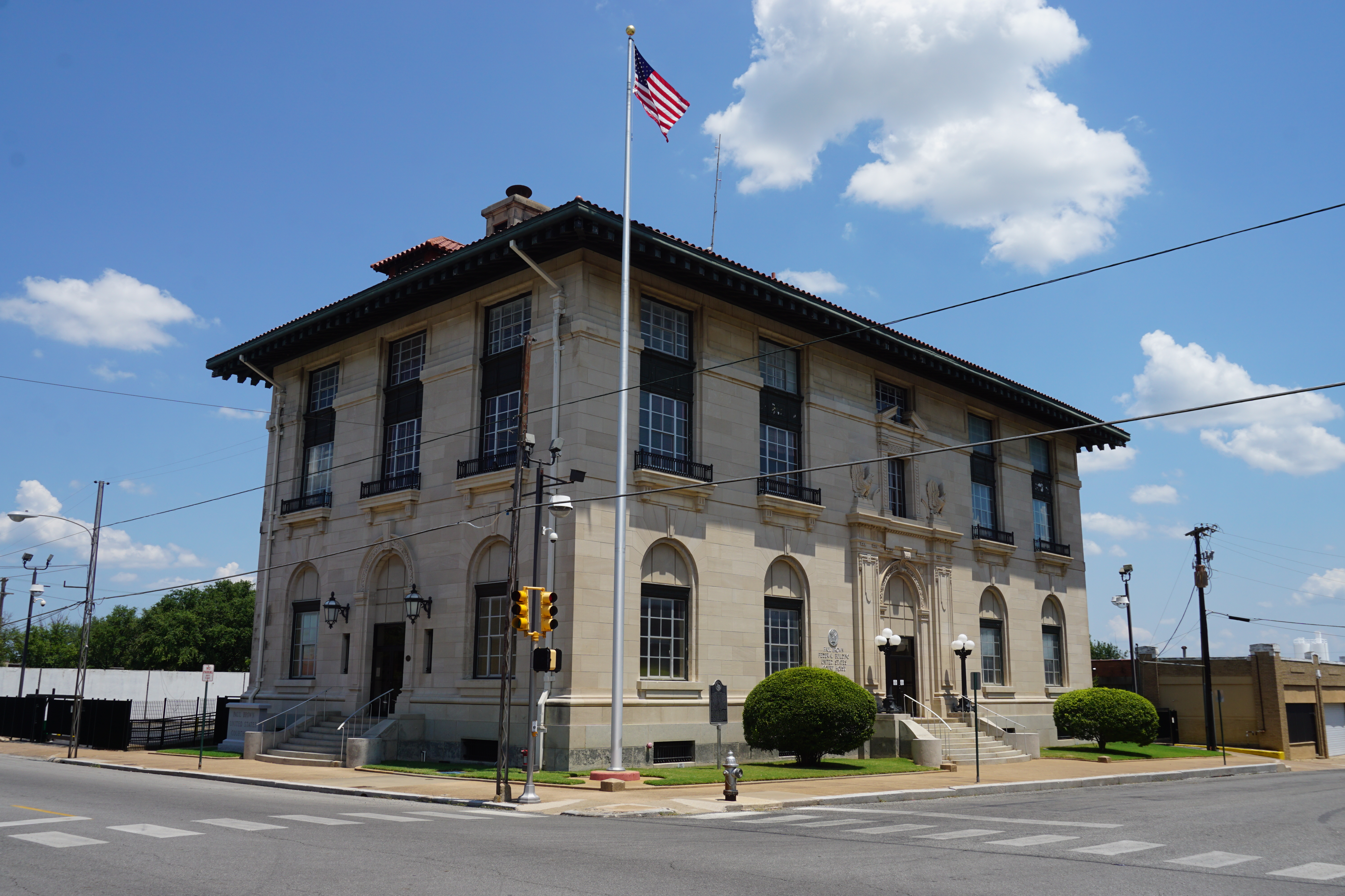 Two-story stone federal building on a street corner with an American flag flying from a tall flagpole.
