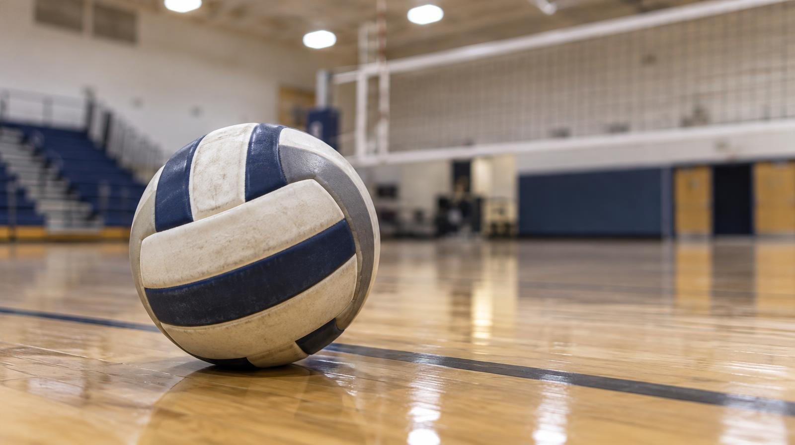 A volleyball resting on a hardwood gym floor near a volleyball net.