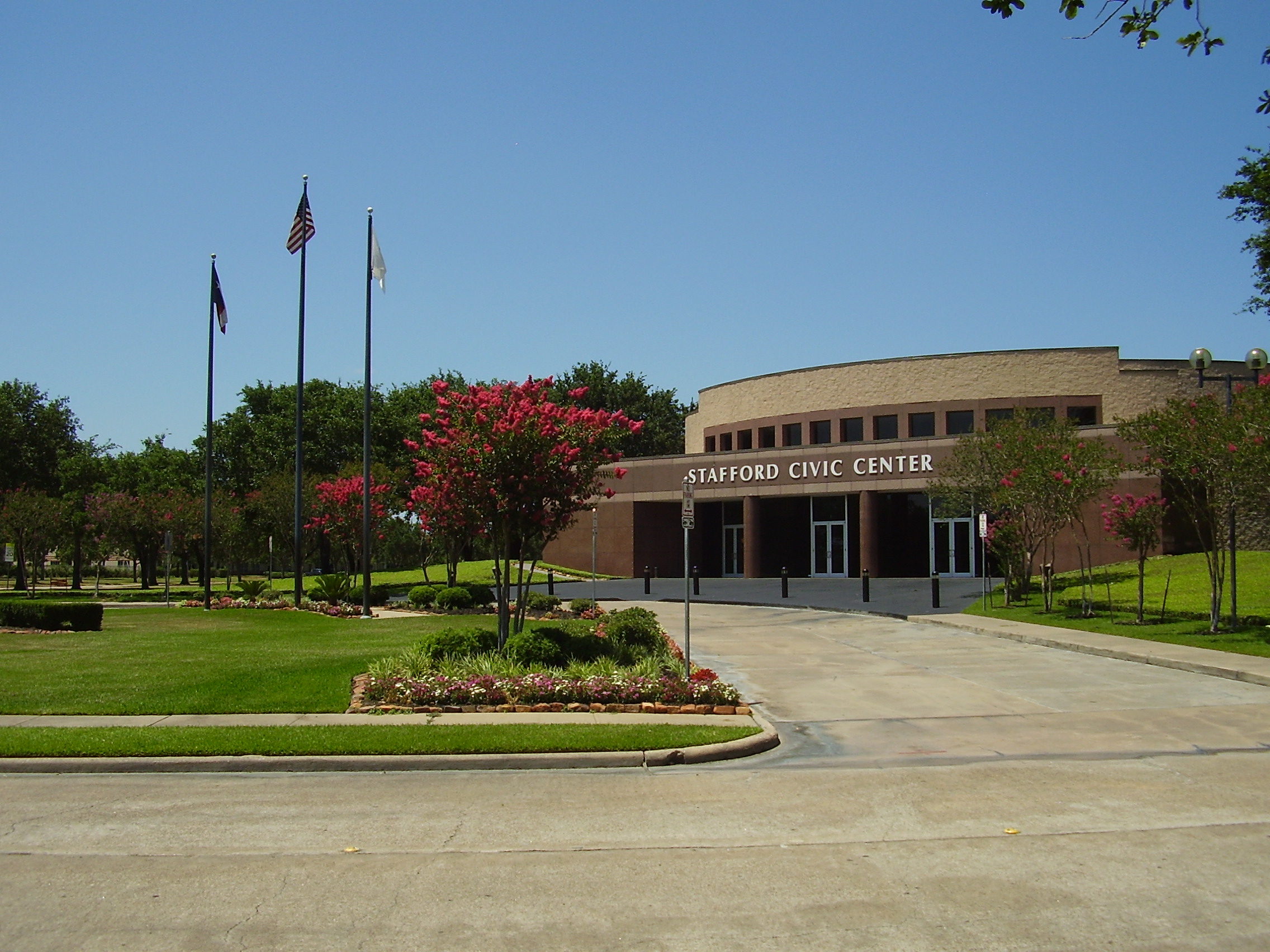 Brick civic center building with three flagpoles and red flowering trees out front.