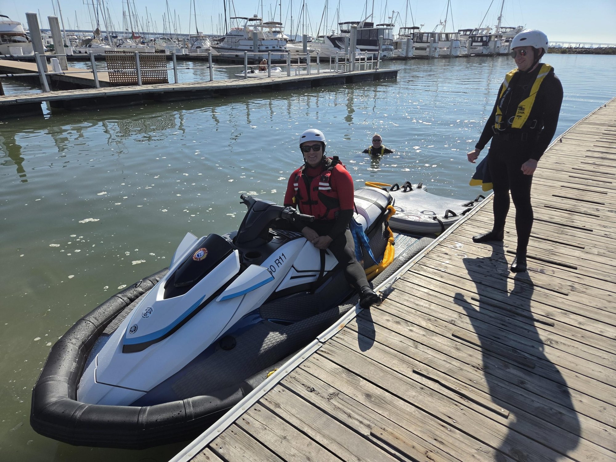 A white and blue WaveRunner labeled 50R11 docked at a marina pier with two people in rescue gear nearby.