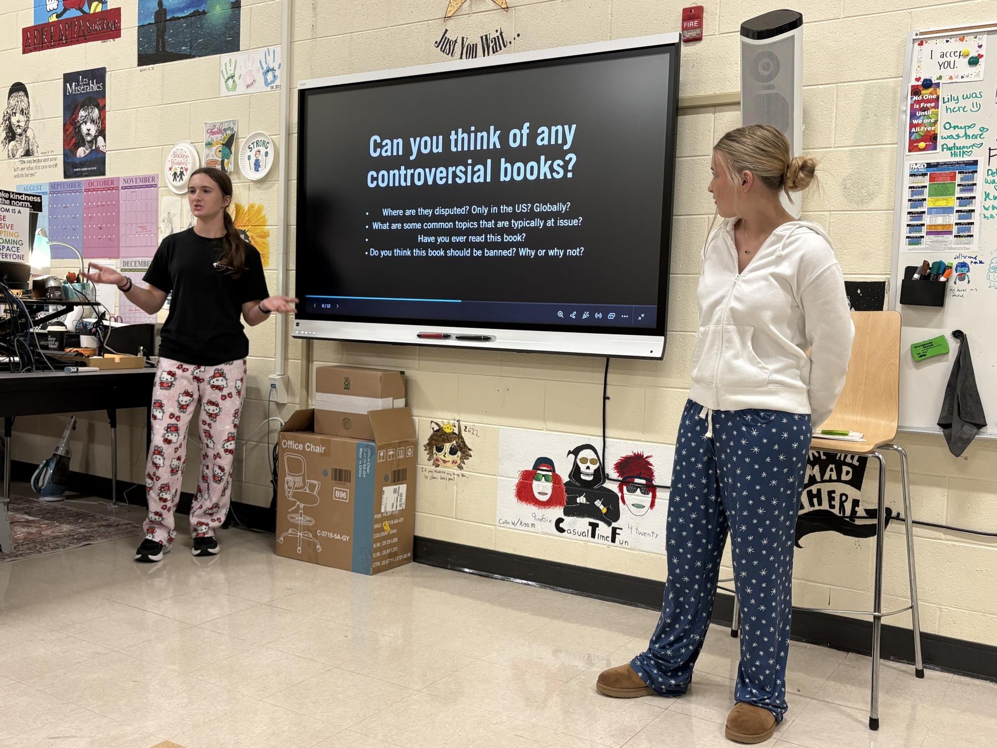 Two students stand before a classroom display screen showing discussion questions about controversial books.