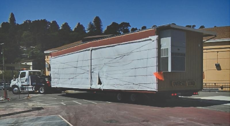 Flatbed truck carrying a large wrapped modular building unit in a parking lot.