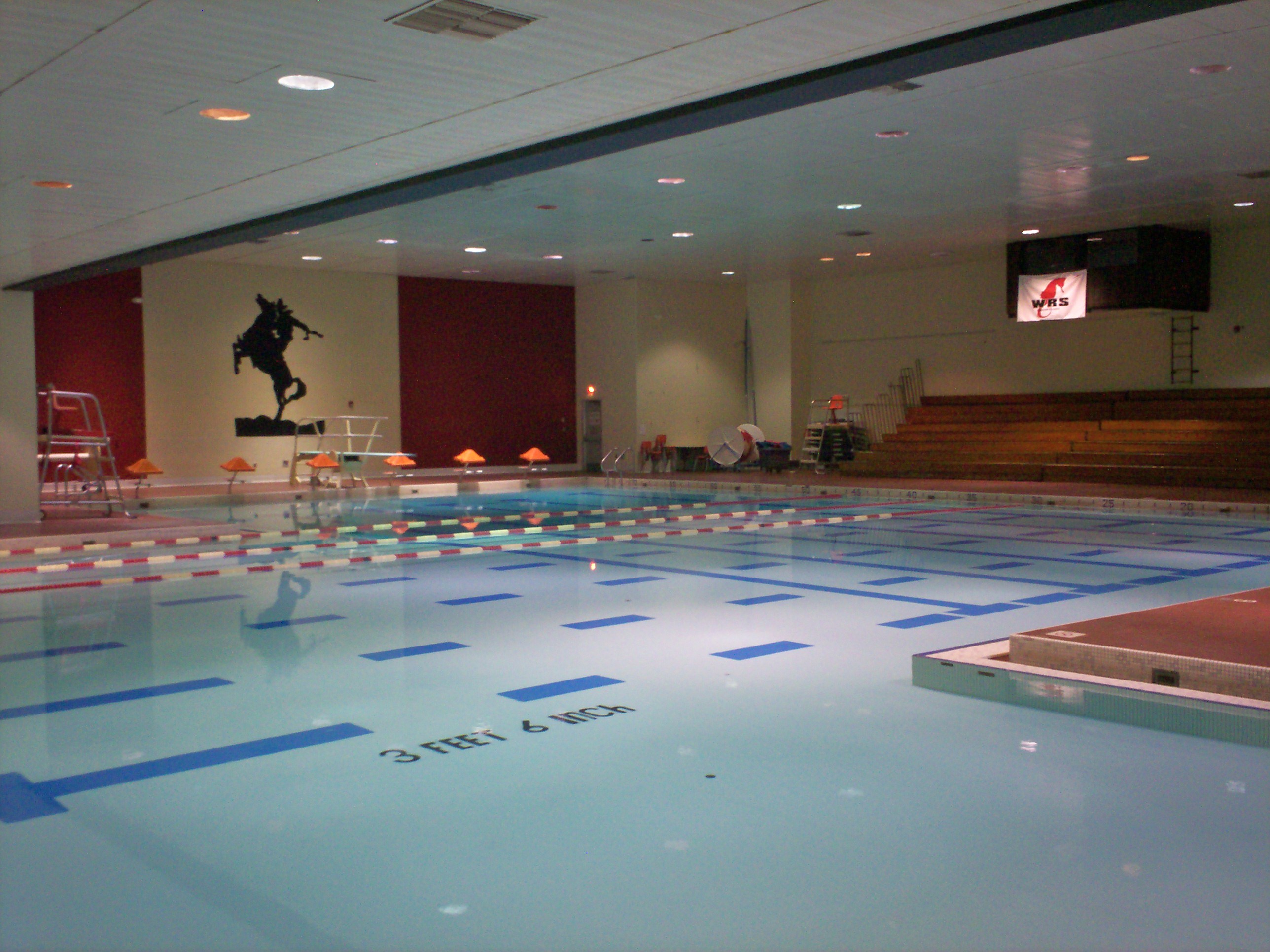 Indoor swimming pool with blue lane dividers, bleachers, and overhead lighting.