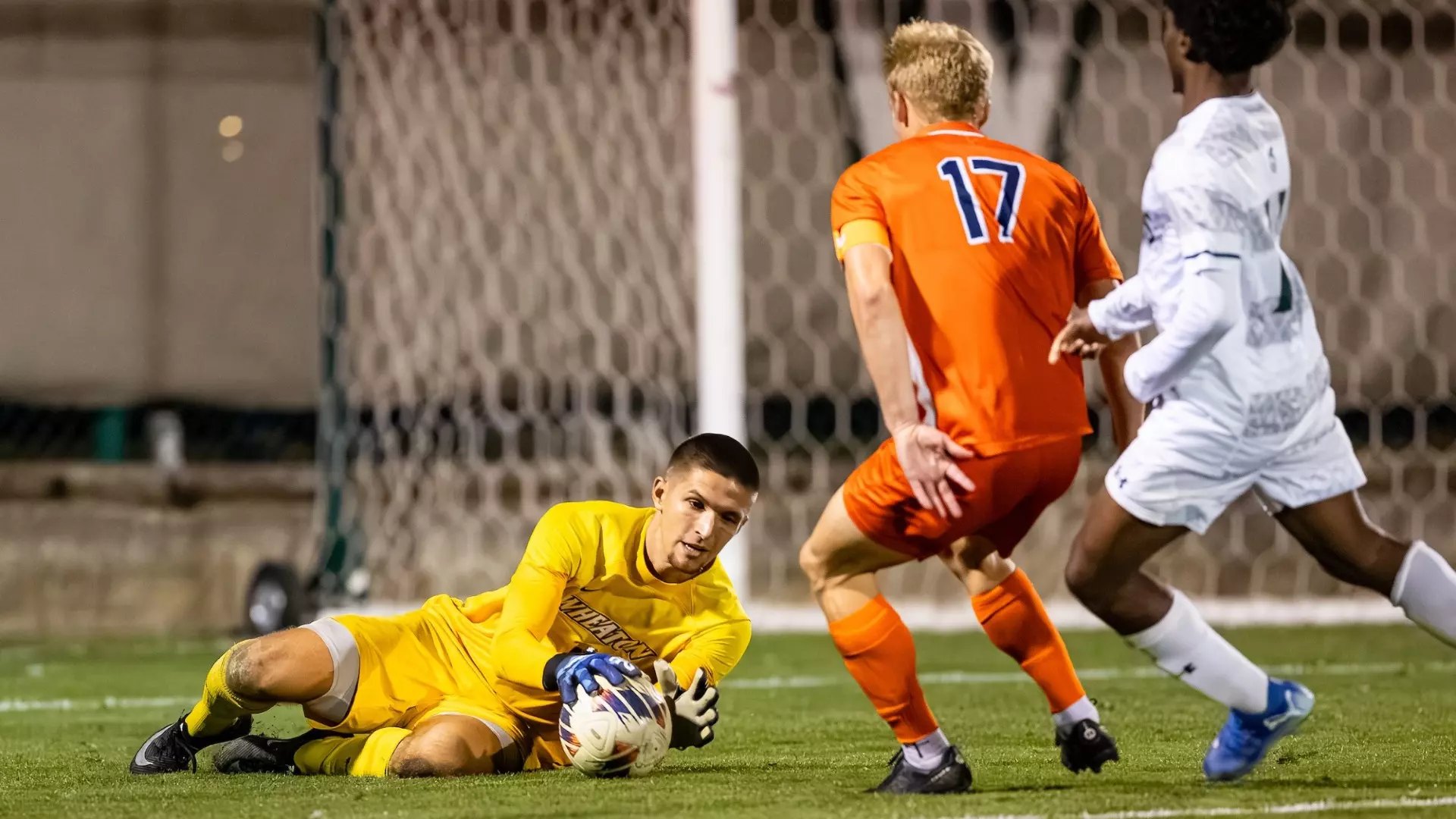 A goalkeeper in a yellow jersey dives to collect the ball on the ground during a nighttime soccer match.