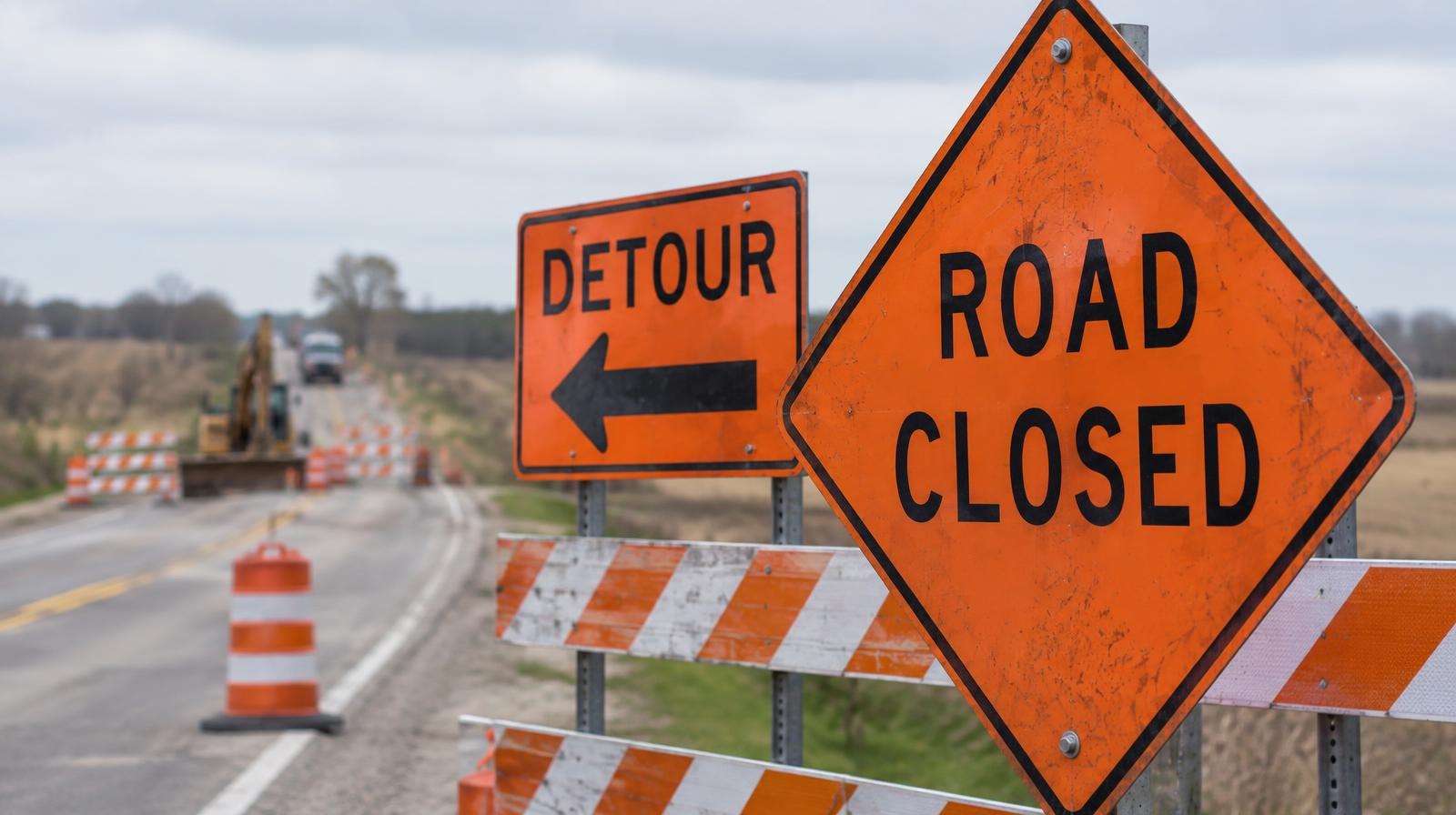 Orange road closure and detour signs with construction barriers on a rural two-lane road.