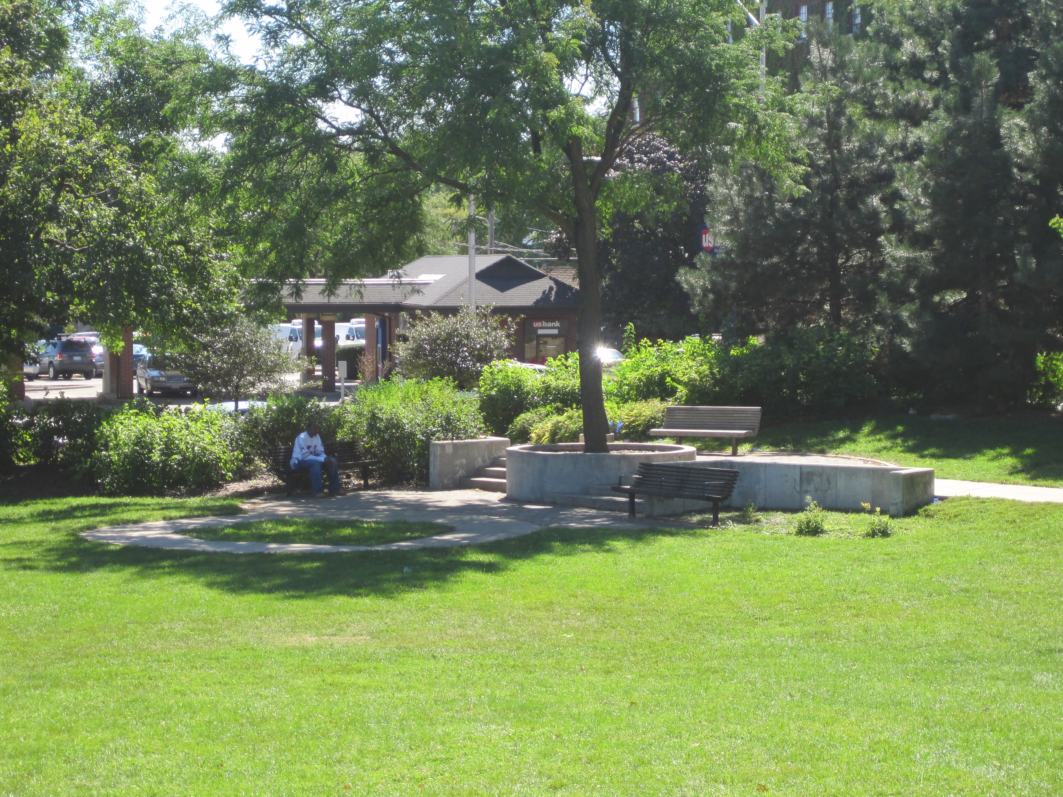 Green lawn with mature trees, benches, and a paved seating area at Scoville Park.