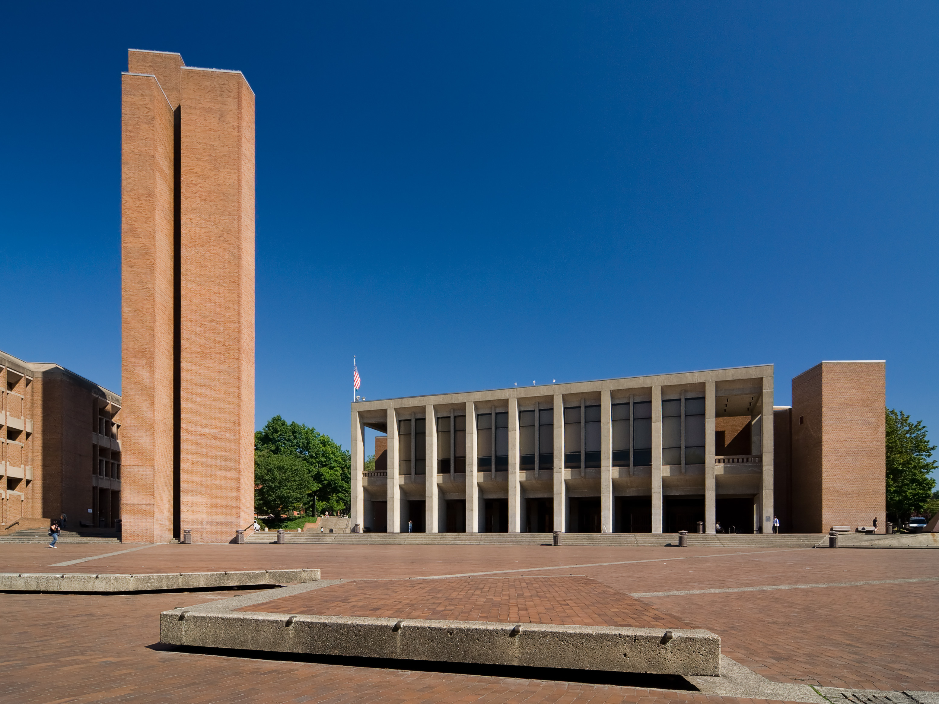 Brick academic building with a tall tower and an American flag on a plaza.