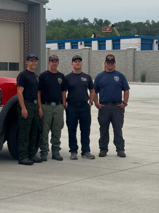 Four firefighters in dark uniforms stand in front of a red fire truck.
