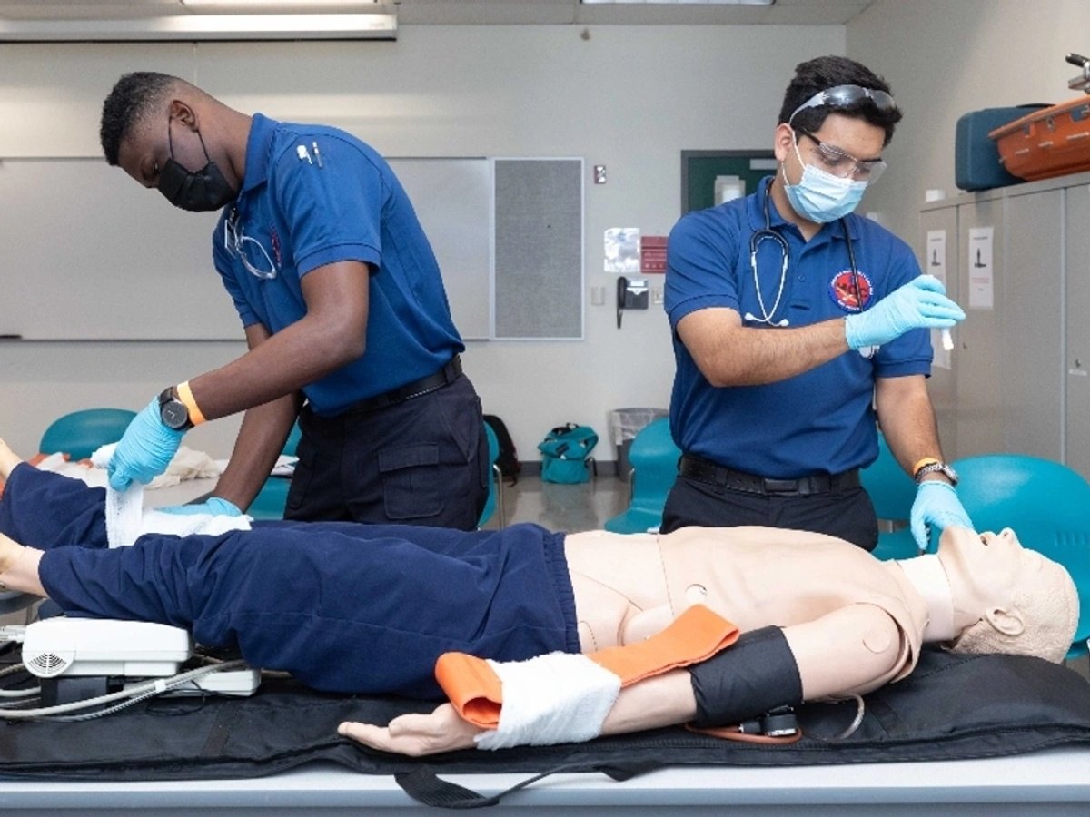 Two students in blue uniforms and masks practice on a medical training mannequin.
