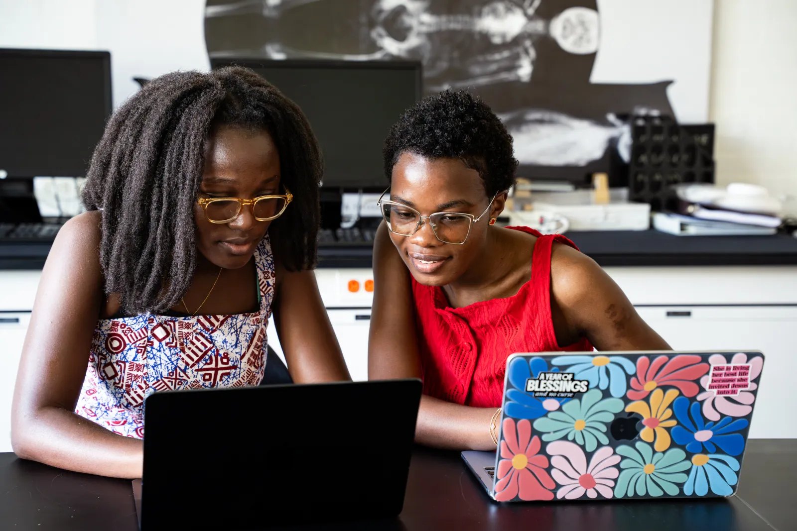 Two women wearing glasses sit together at a table, looking at a laptop screen.