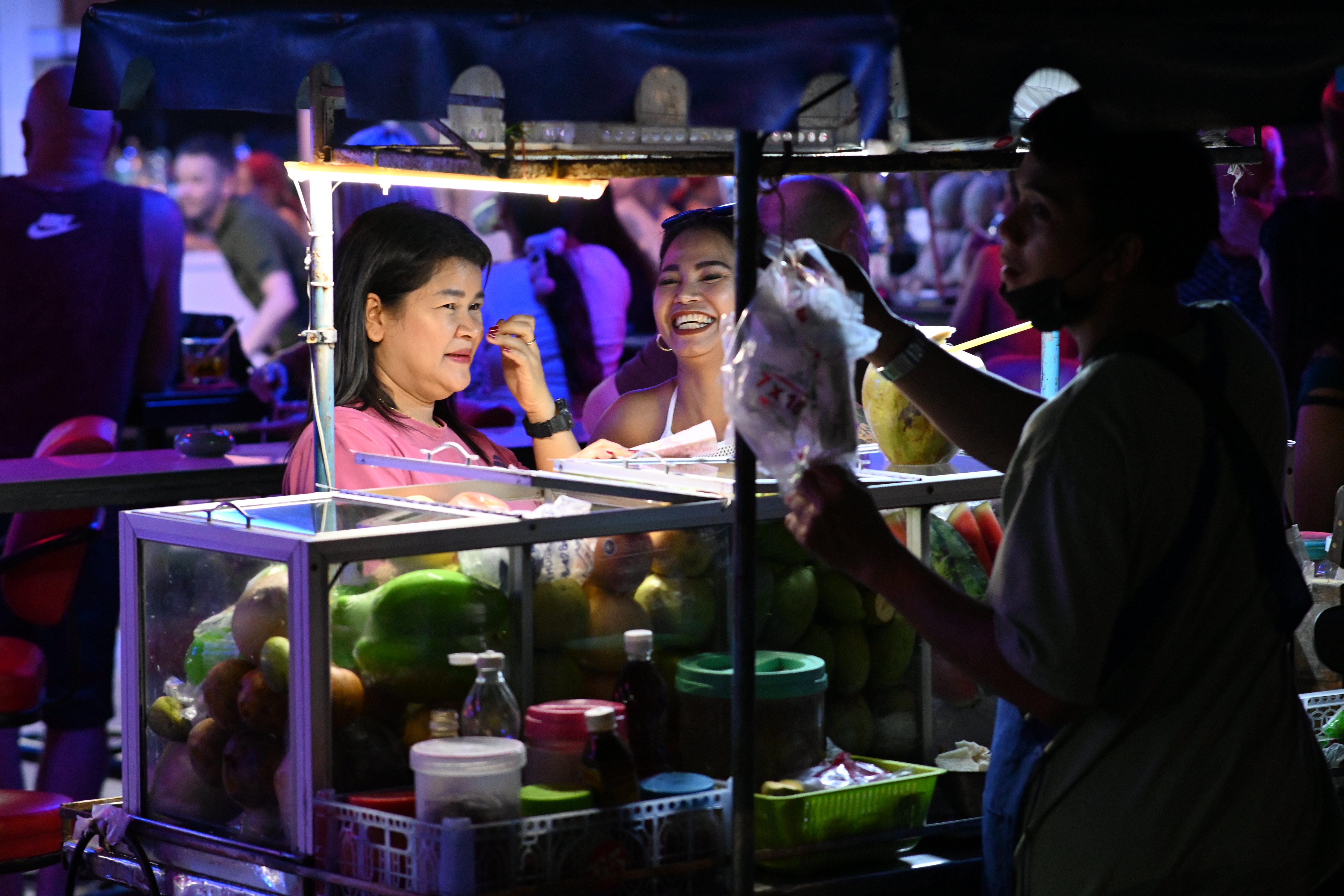 Smiling vendors stand behind a brightly lit food cart at a festive outdoor market.