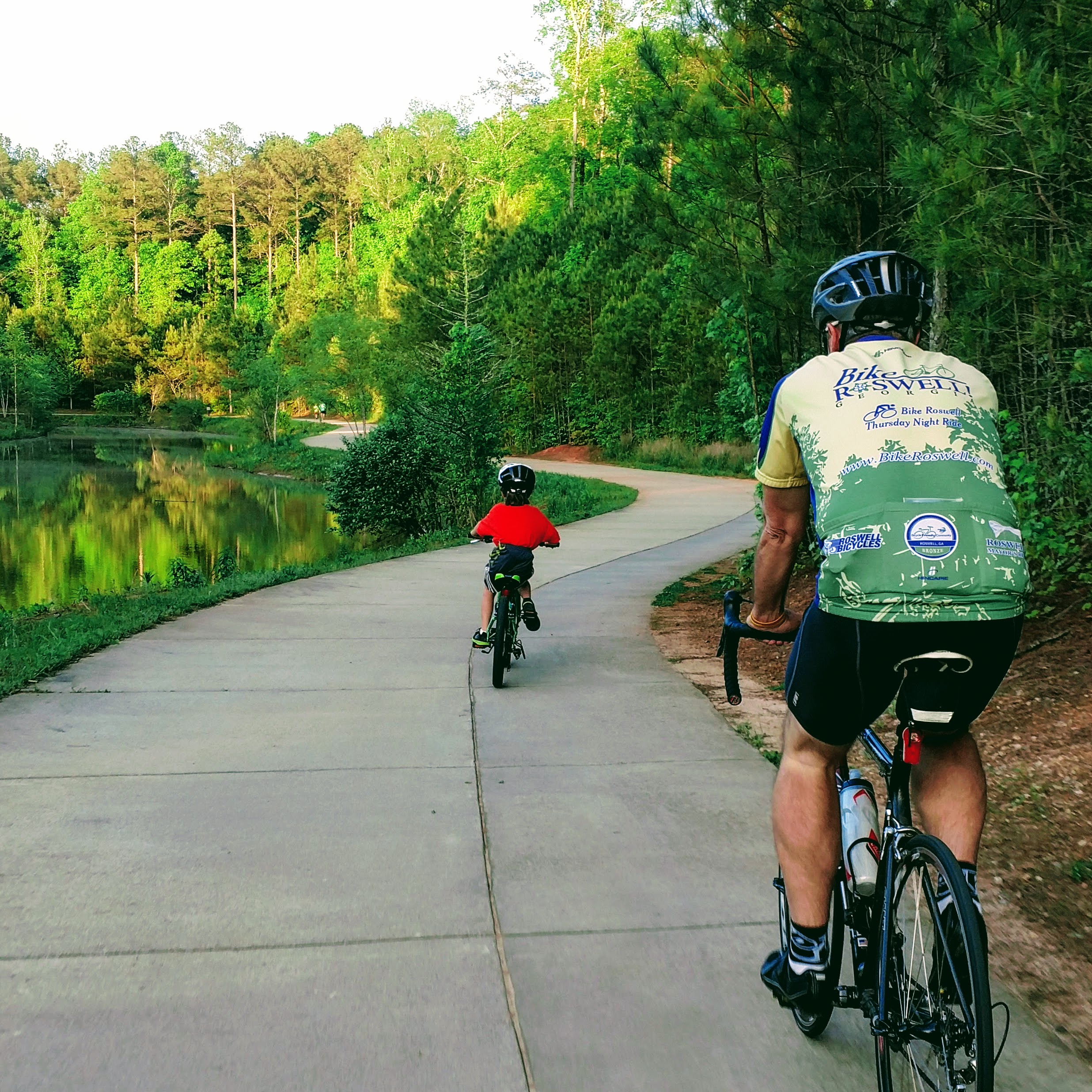 Adult and child cyclists riding on a paved greenway trail alongside a pond.