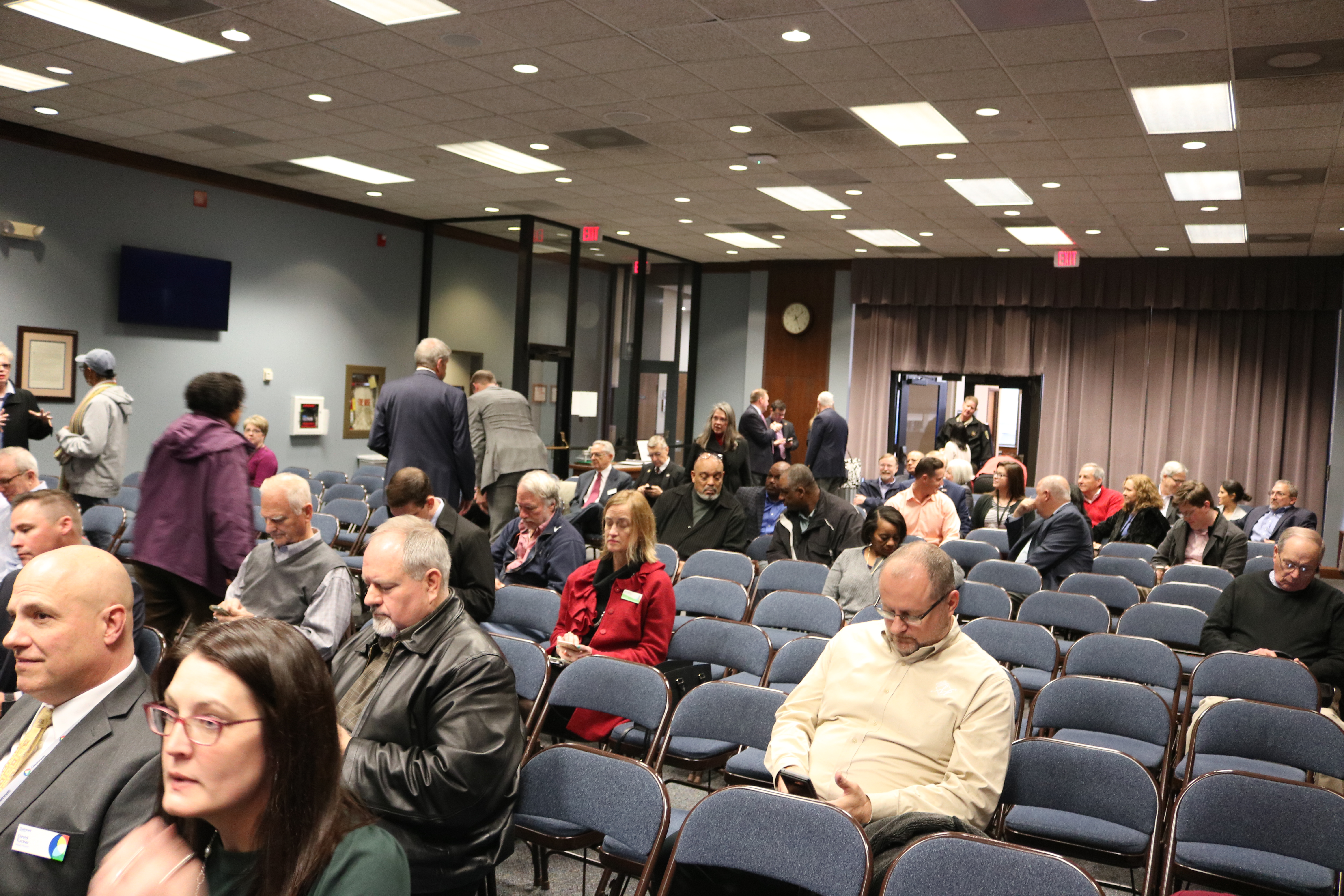 Crowded government meeting room with rows of blue chairs and many attendees seated and standing.