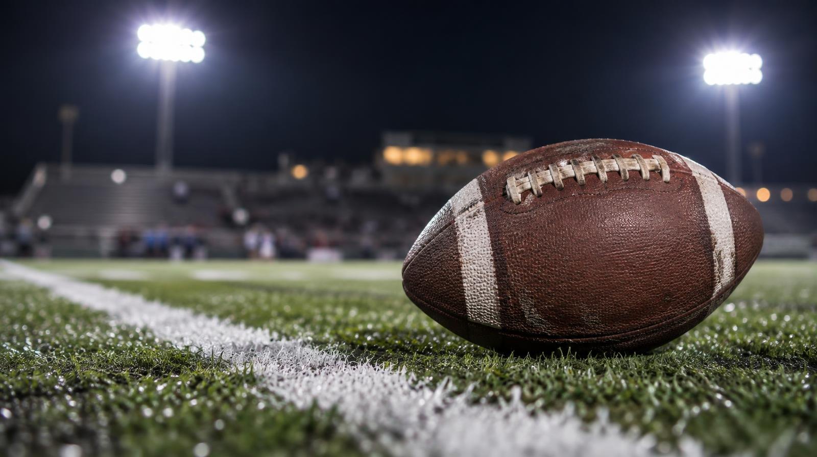 An American football on green artificial turf beside a white yard line under bright lights.