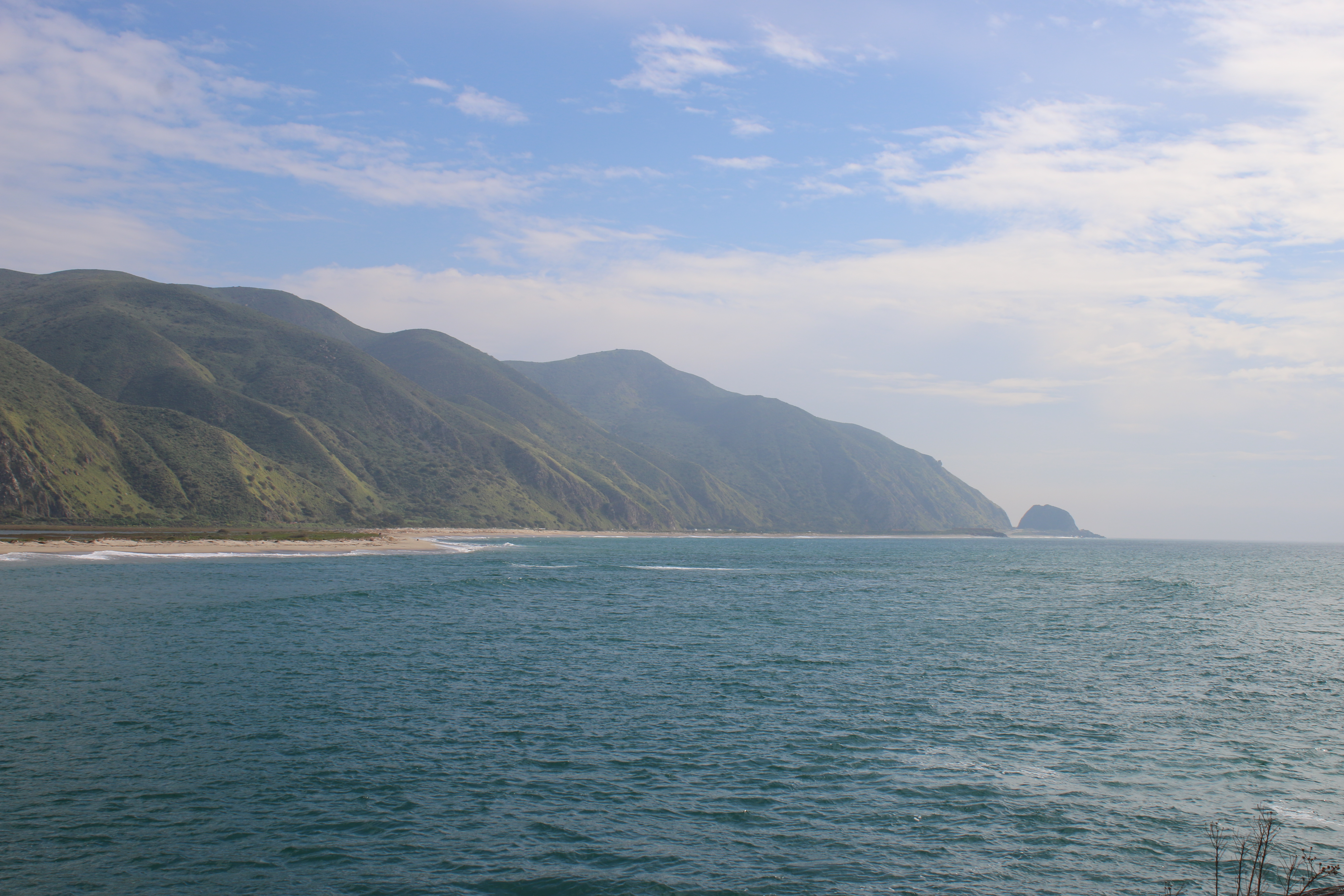 Coastal lagoon with green hills, sandy beach, and a large rock formation in the distance.