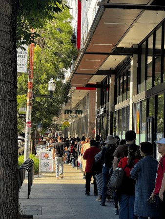 A long line of people stretches down a city sidewalk outside a building during daytime.