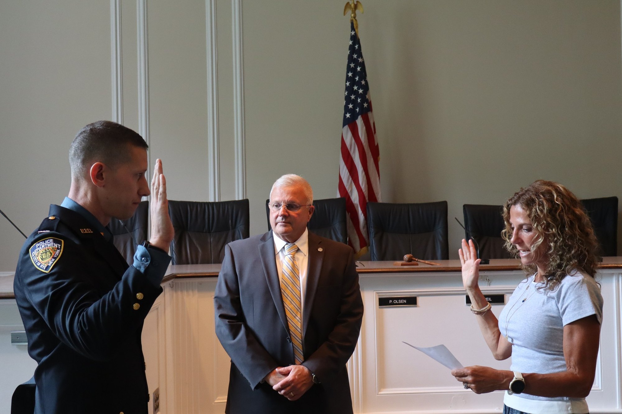 Three people in a formal chamber room during a swearing-in ceremony, one in police uniform with hand raised.