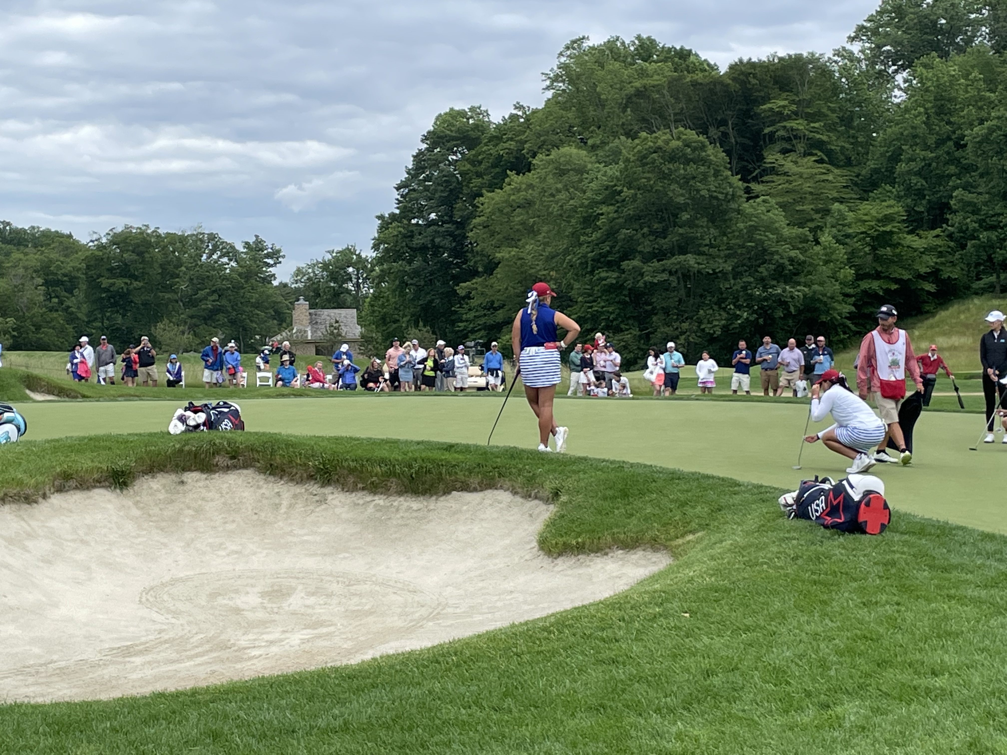 Golfers on a putting green at a golf club with a sand bunker and spectators in the background.