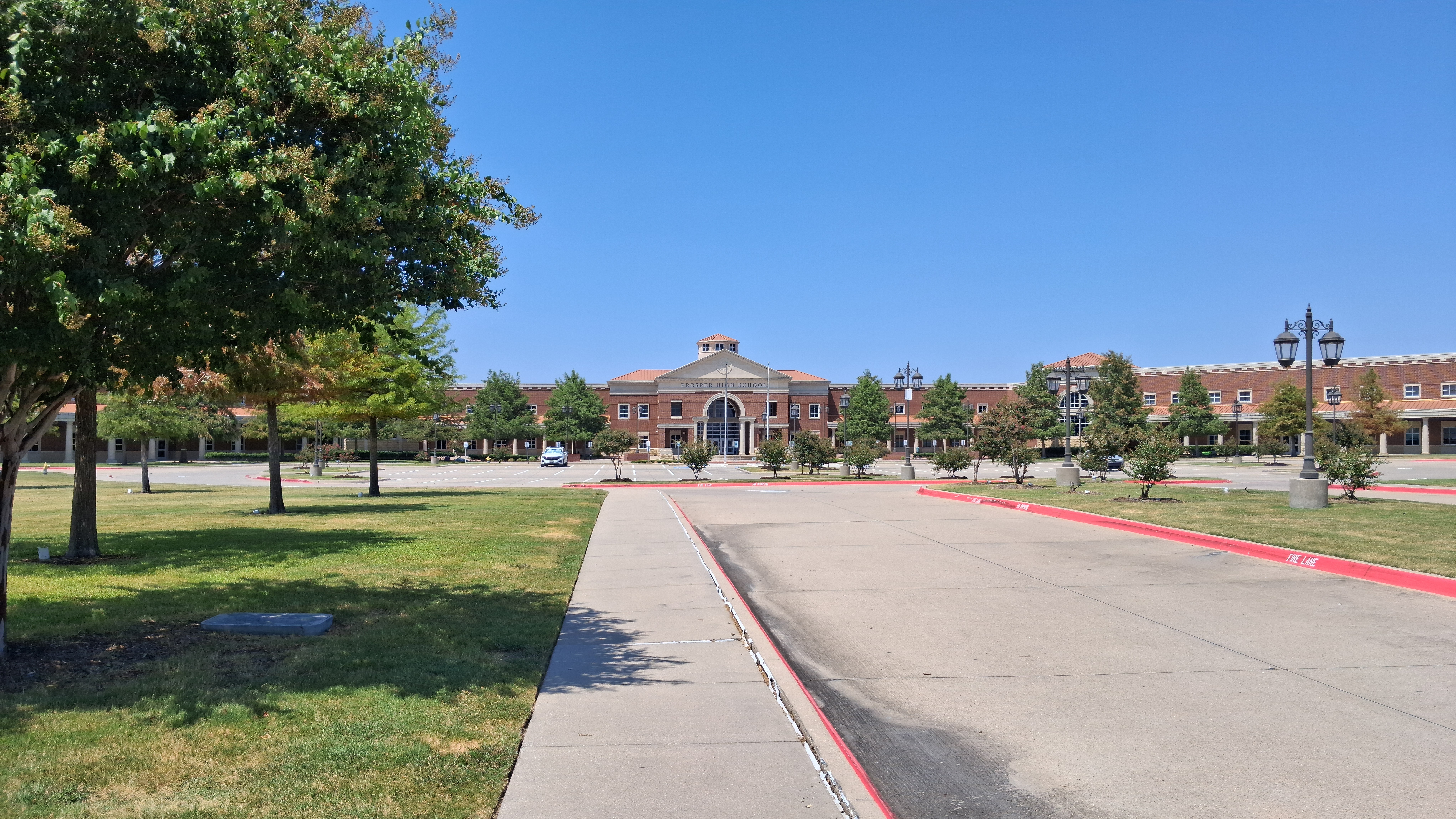 Brick school building entrance with circular drive, lawn, and lamp posts under blue sky.