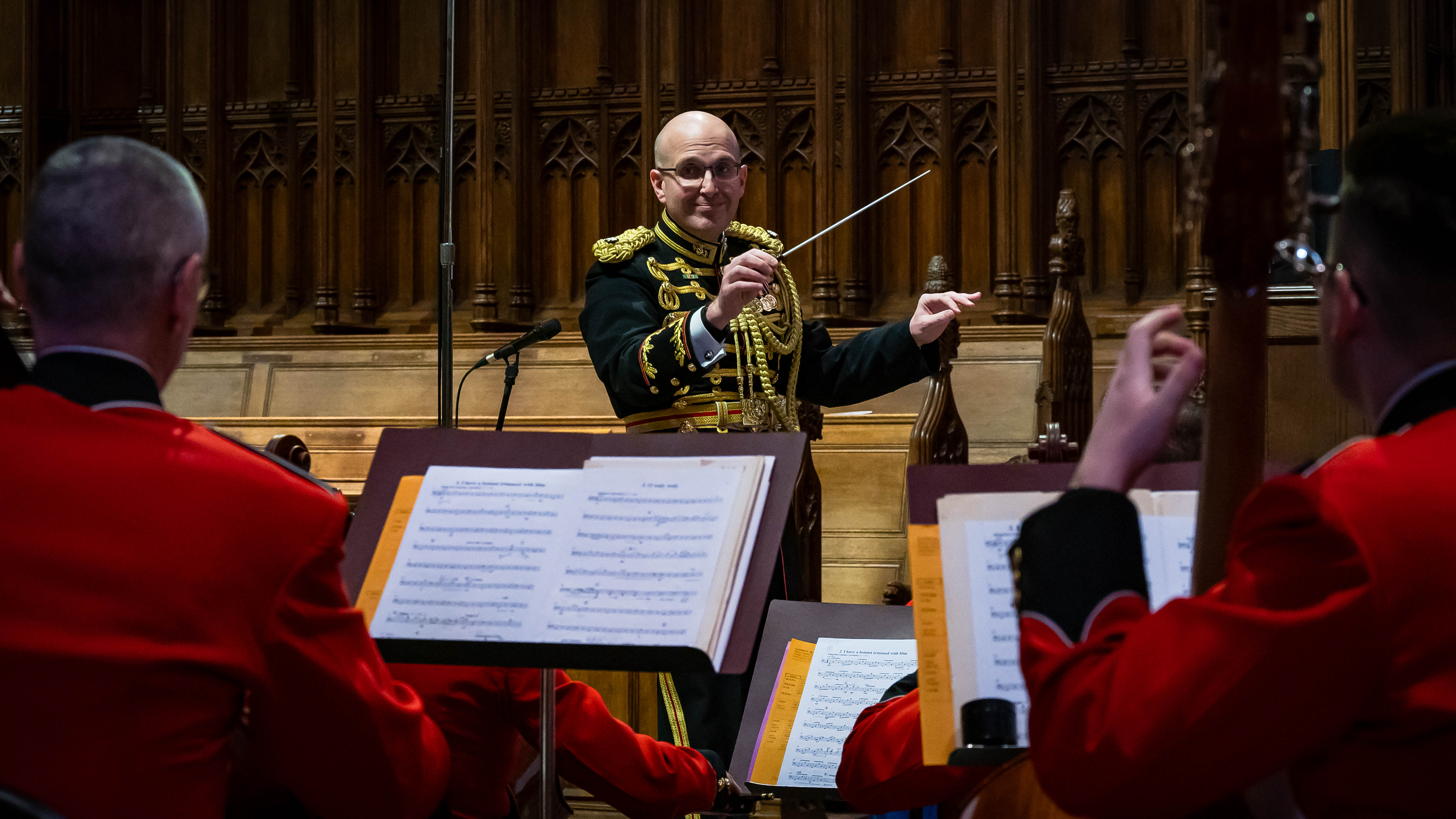 Military conductor in dark uniform raises baton before musicians in red dress uniforms.