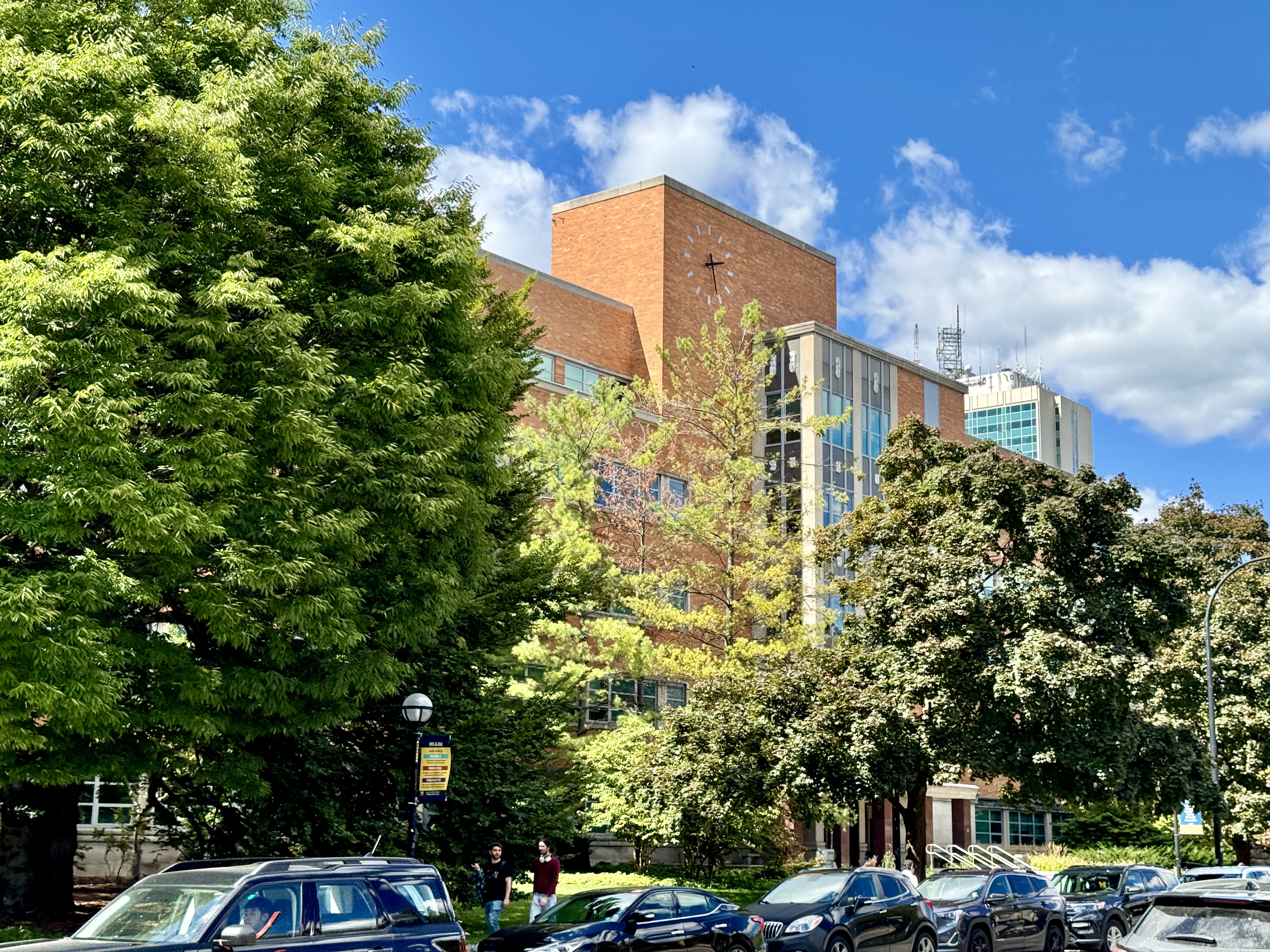 Brick mid-century university building partially obscured by trees, with parked cars in the foreground.