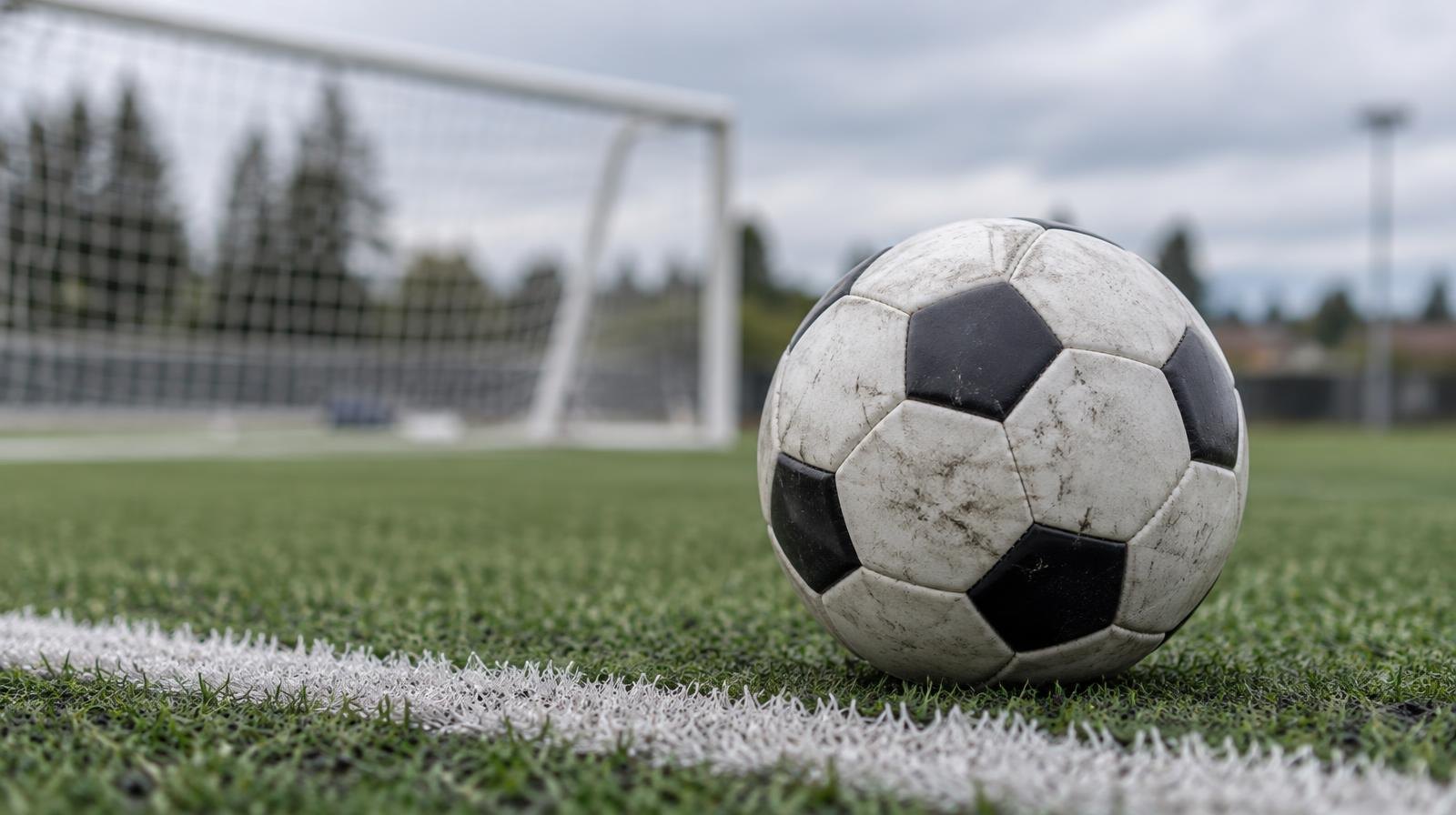 A soccer ball resting on green artificial turf beside a white yard line with a goal net in the background.