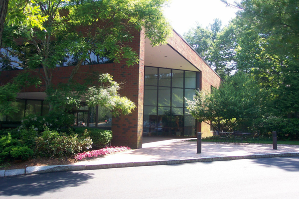A two-story red brick office building with large glass windows surrounded by trees and landscaping.
