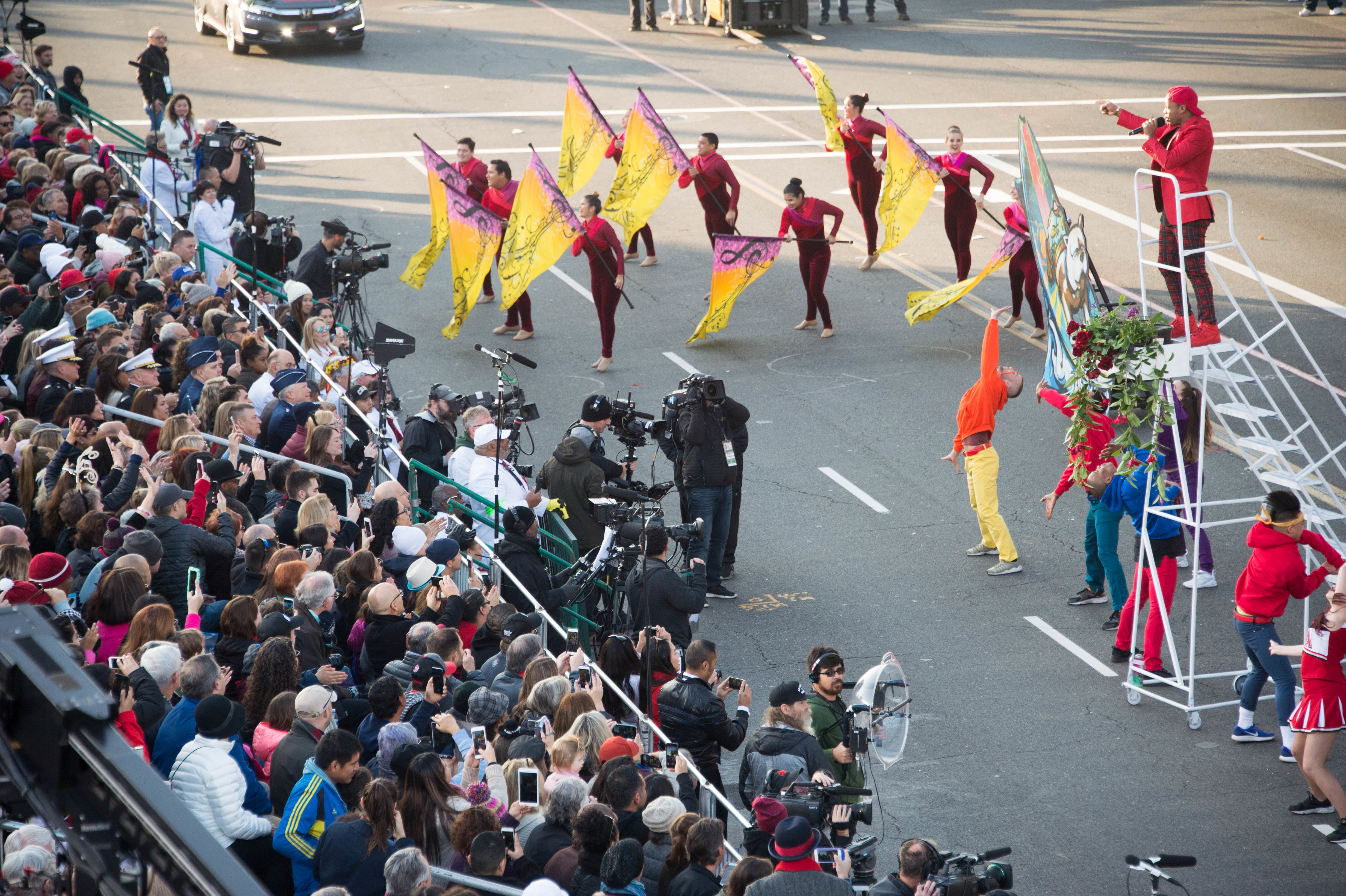 Performers and flag bearers march along a parade route lined with spectators.