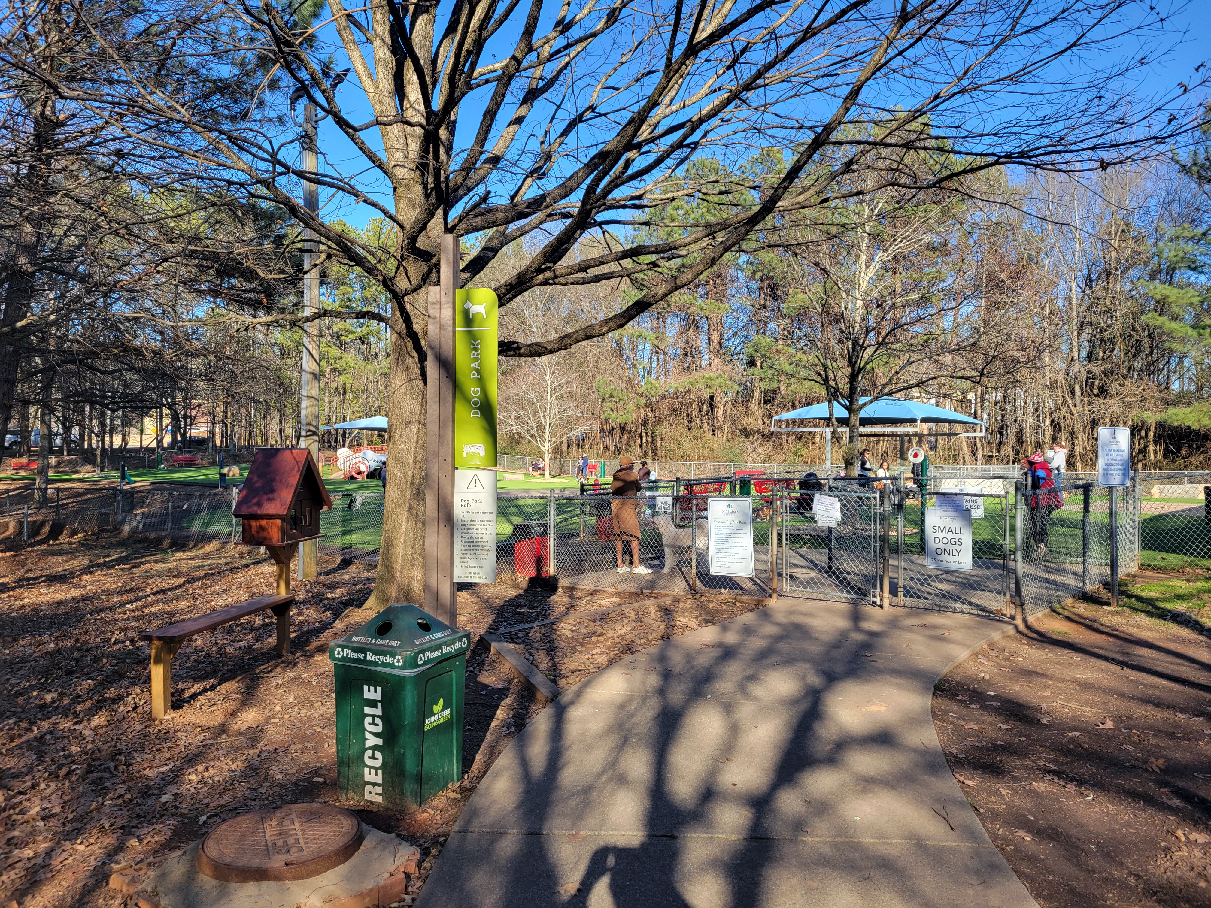Fenced dog park entrance with a paved path, chain-link fence, and bare trees.