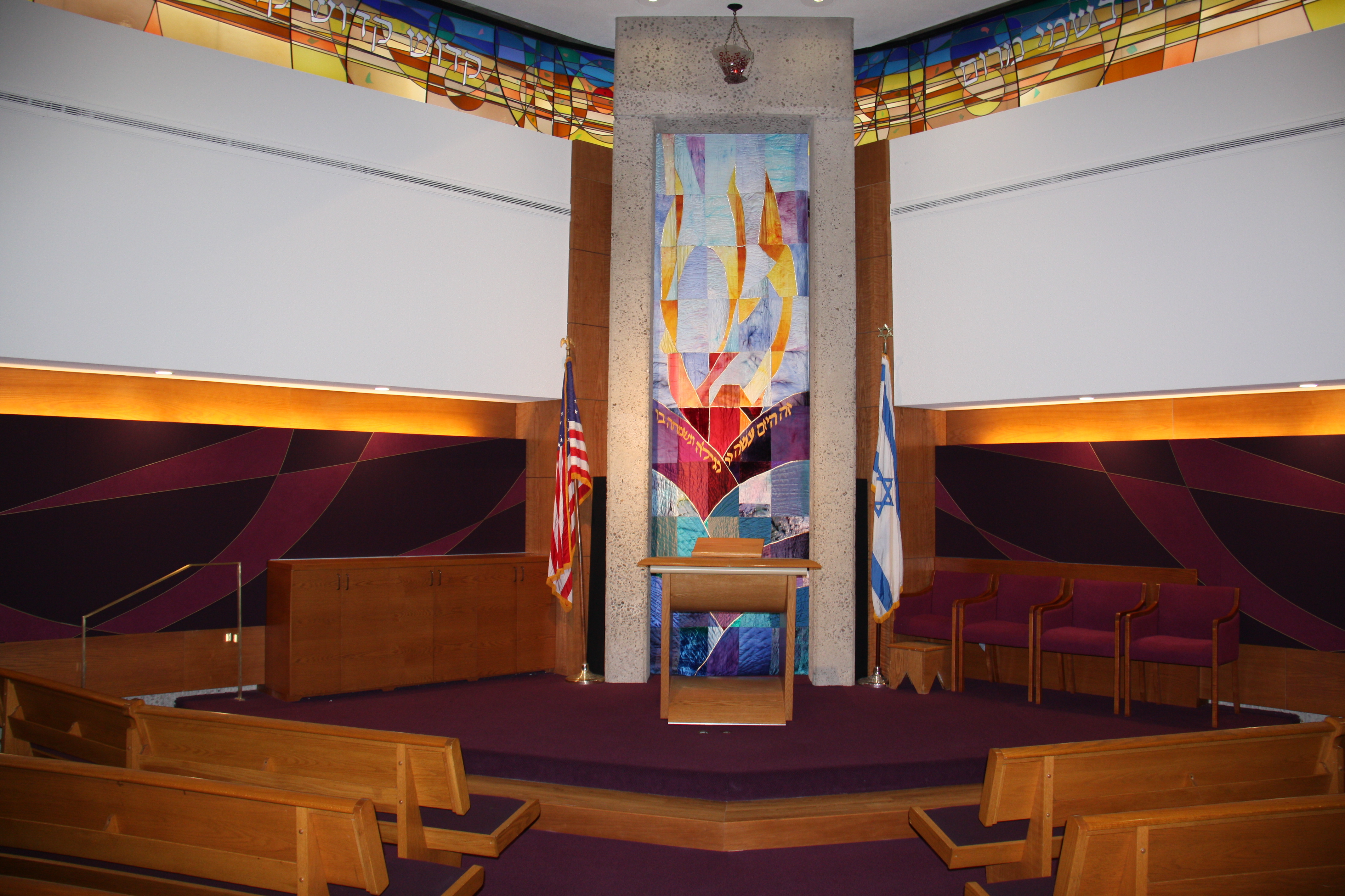 Synagogue sanctuary interior with stained-glass ark, podium, and rows of pews.