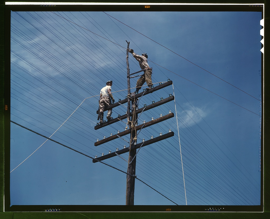 Two workers high on a wooden utility pole with crossarms and power lines against a blue sky.