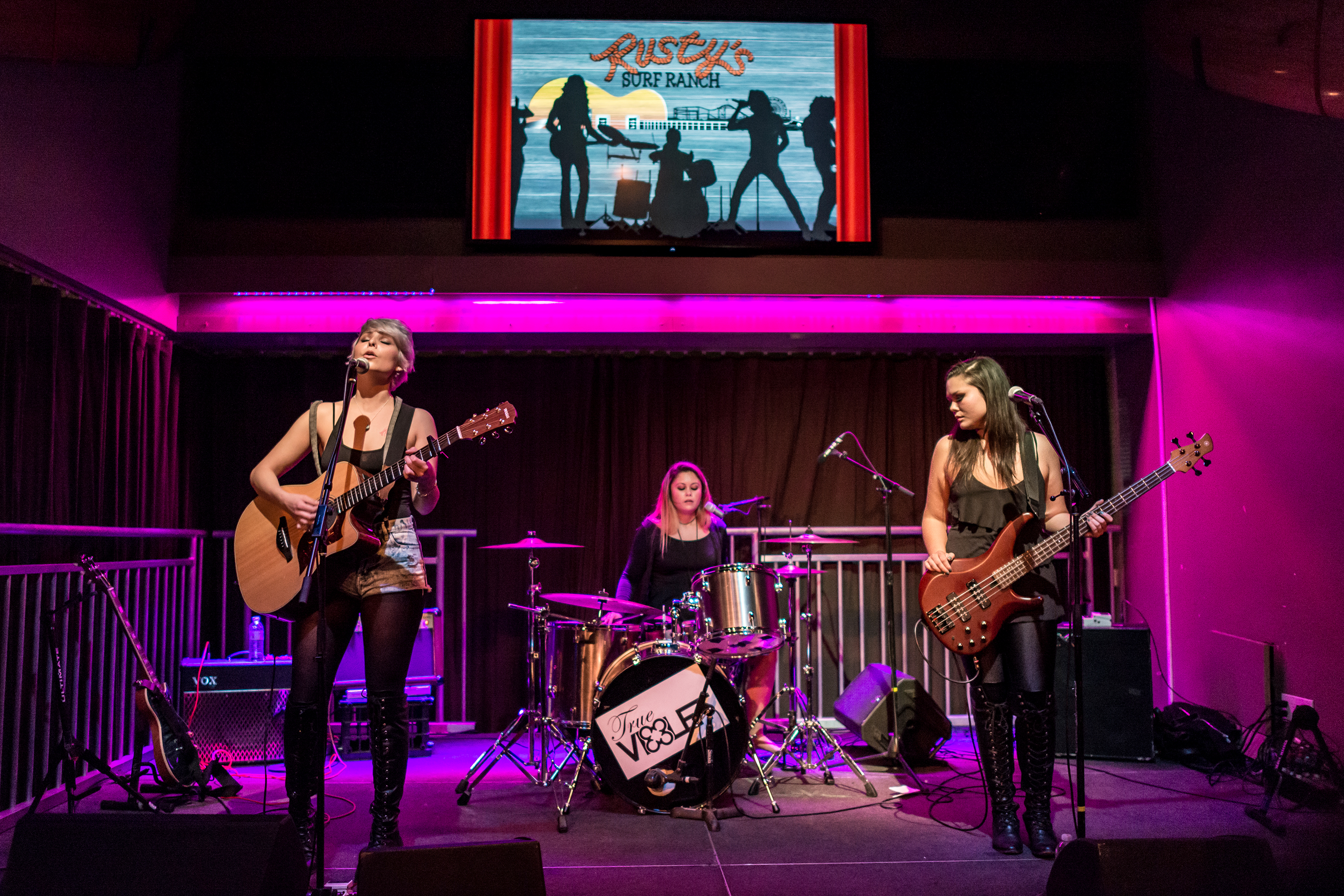 Three musicians performing on a purple-lit stage with a venue logo projected above them.