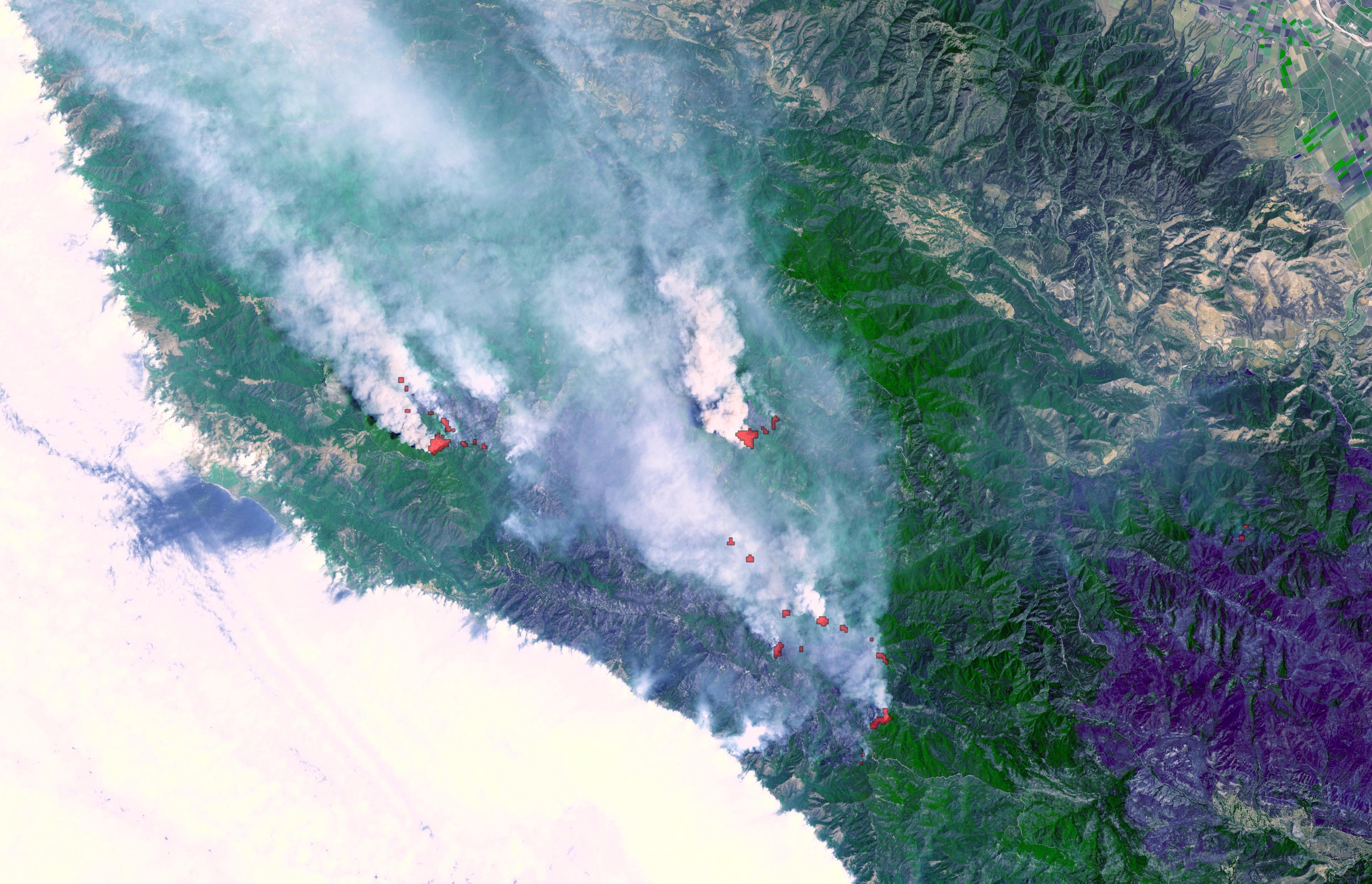 Satellite view of wildfires burning near a coastline with white smoke plumes rising.