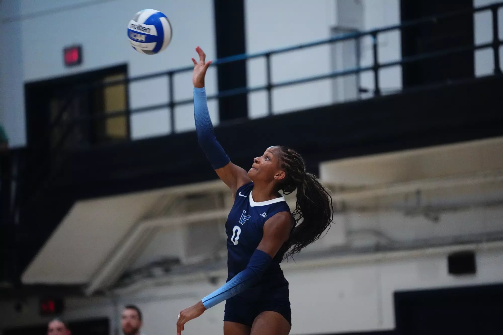 A female volleyball player in a navy uniform jumps to spike a volleyball indoors.