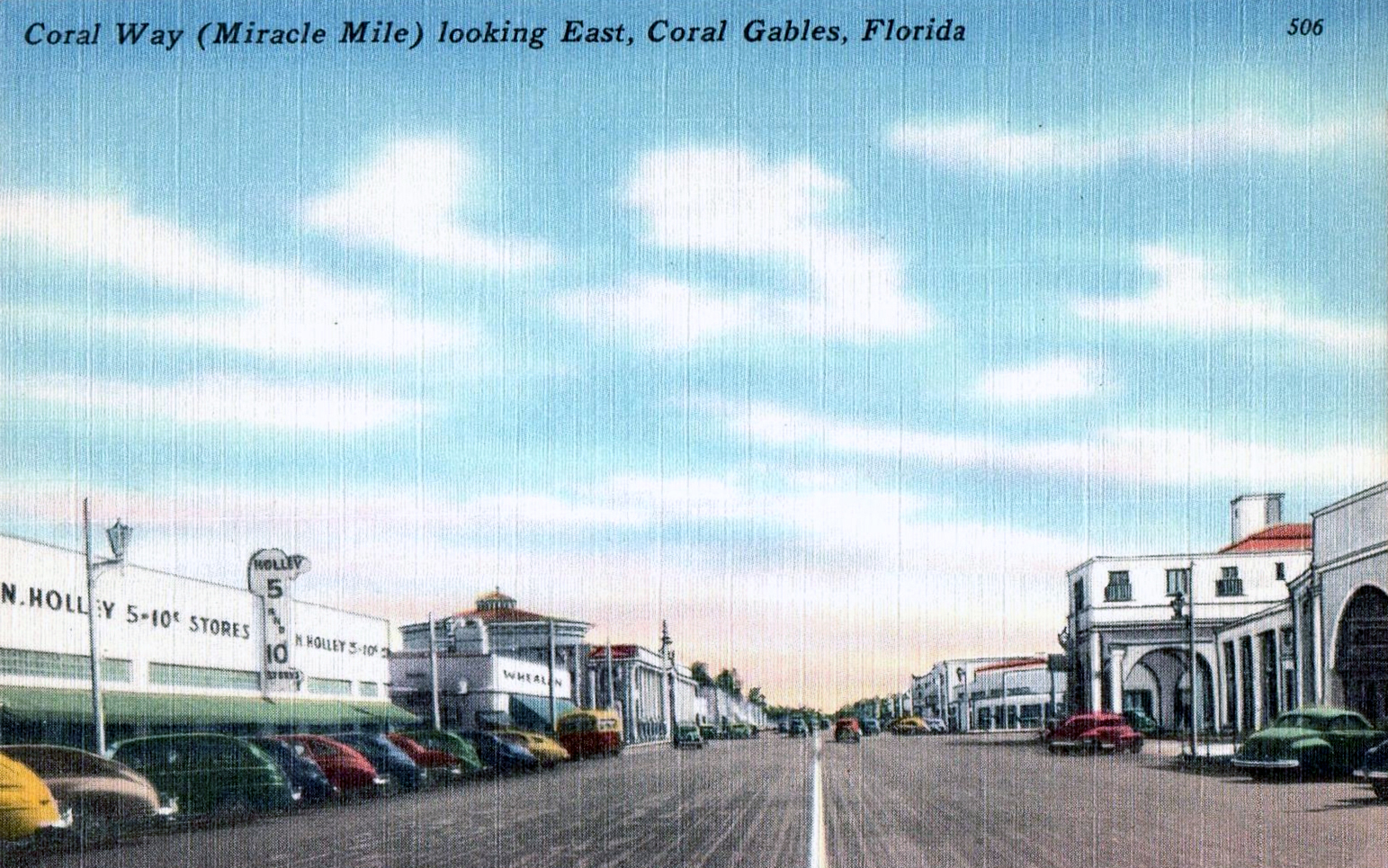 Vintage colorized postcard of a wide commercial boulevard lined with storefronts and parked cars.
