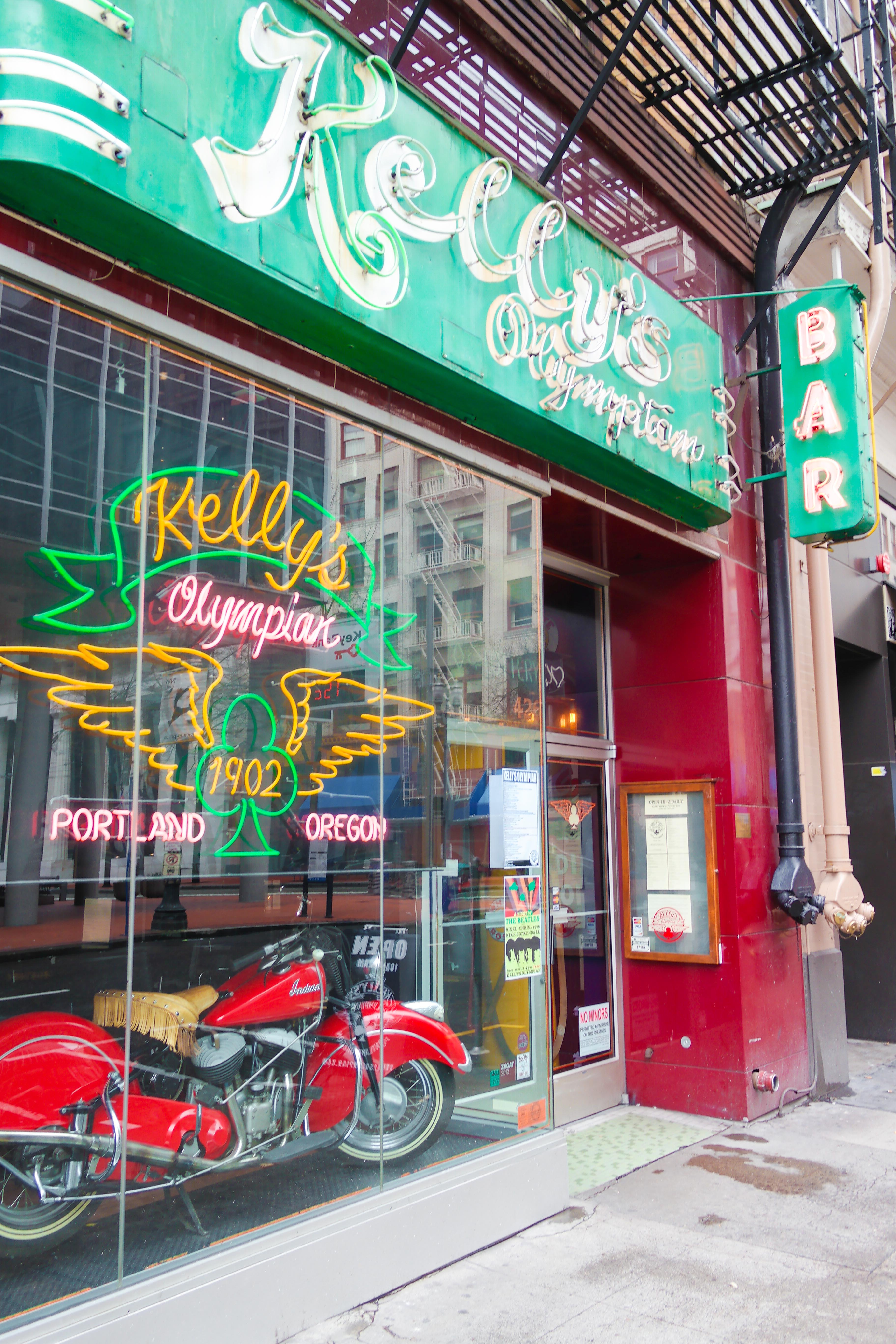 Storefront of a bar with green neon sign and vintage motorcycle visible in window.