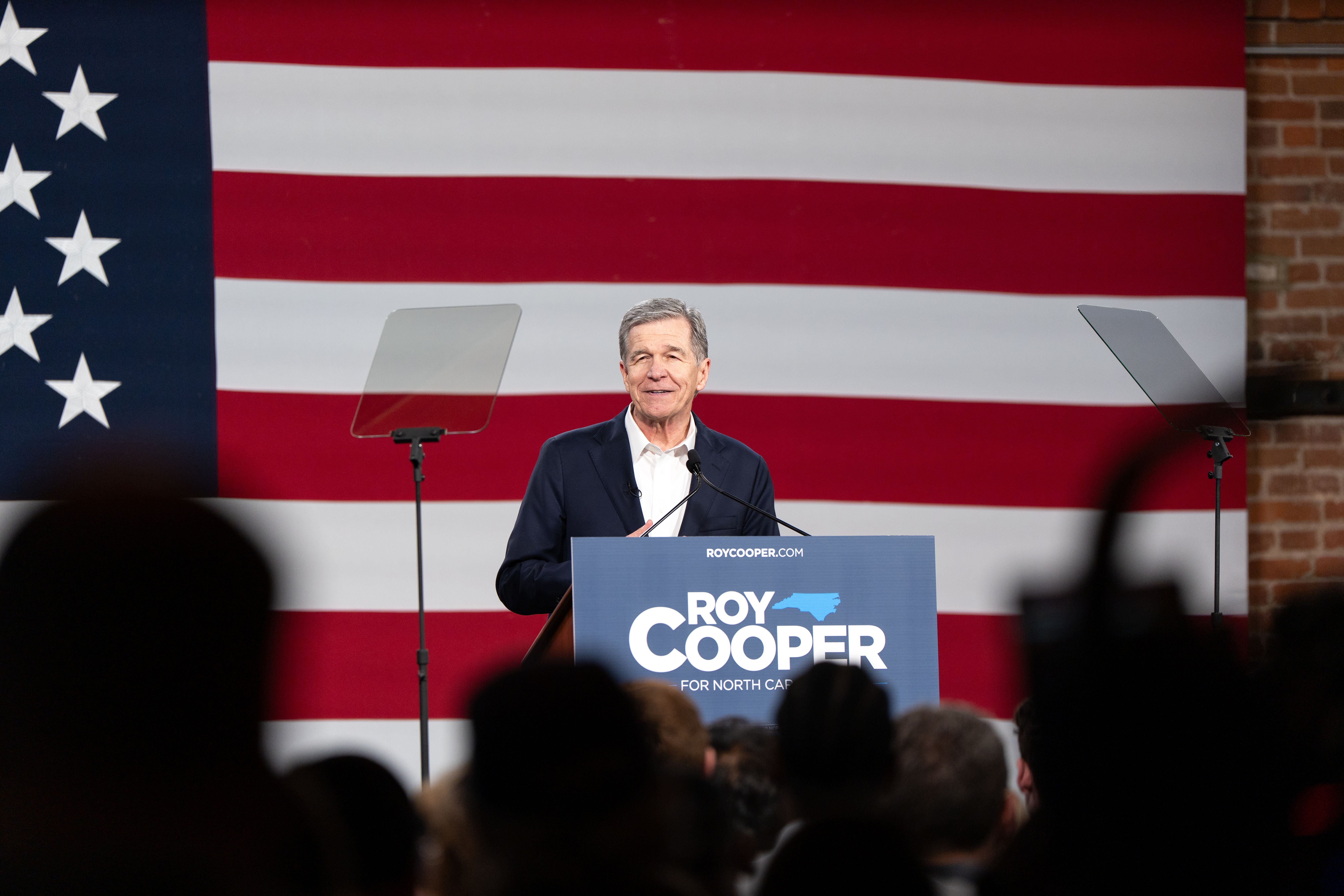 Man in dark blazer speaks at a podium with a campaign sign before a large American flag.