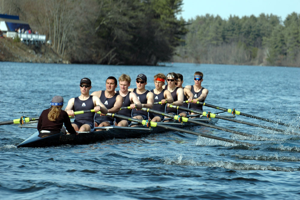 Eight rowers and a coxswain race in a shell on a calm river in early spring.